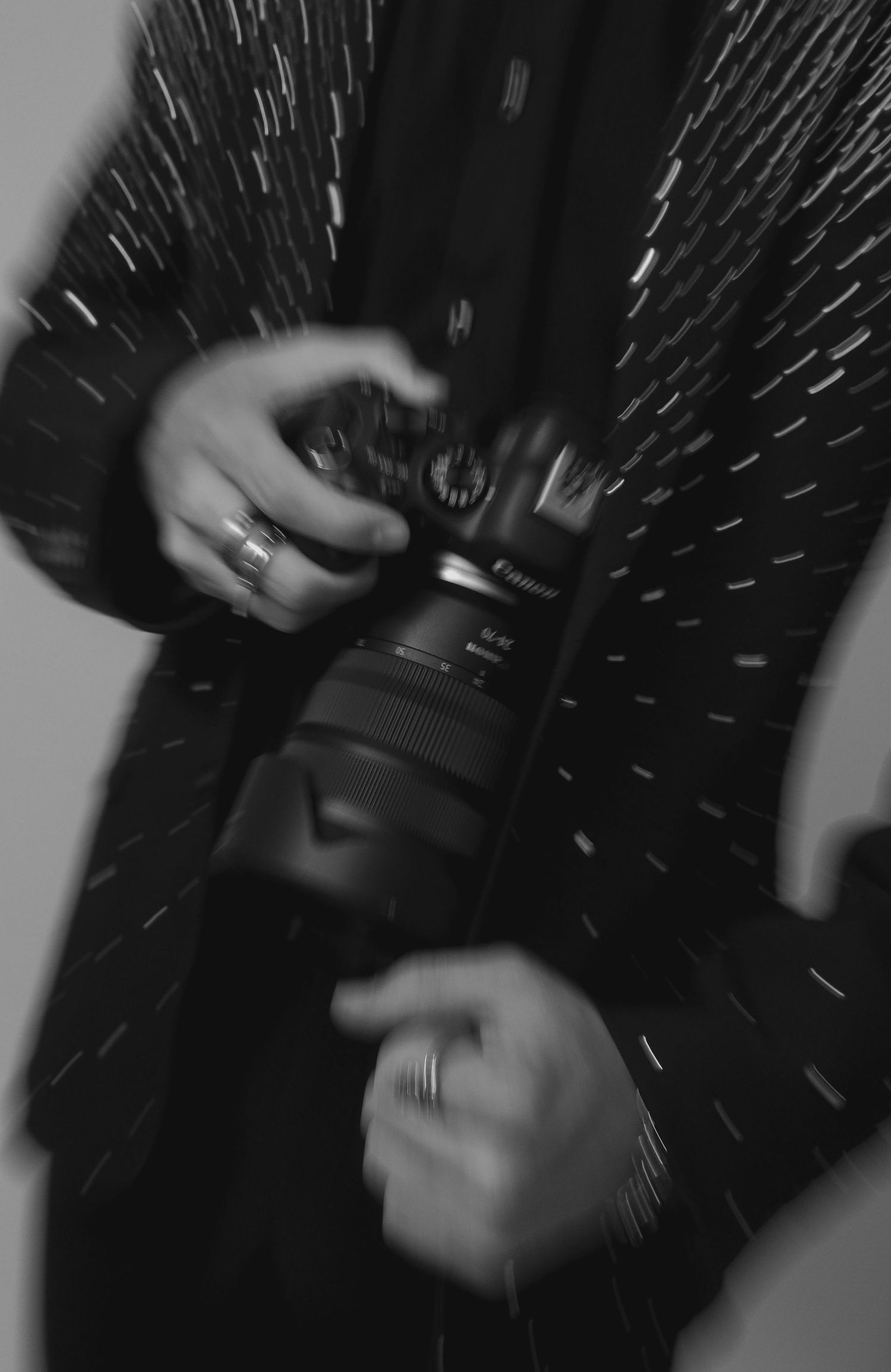 Black and white photo of a person holding a Canon camera with a zoom lens, wearing rings, with a starburst light effect.