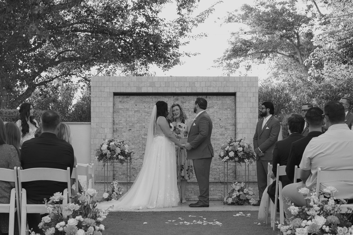 Black and white outdoor wedding ceremony with bride and groom at floral altar, guests seated in white chairs.