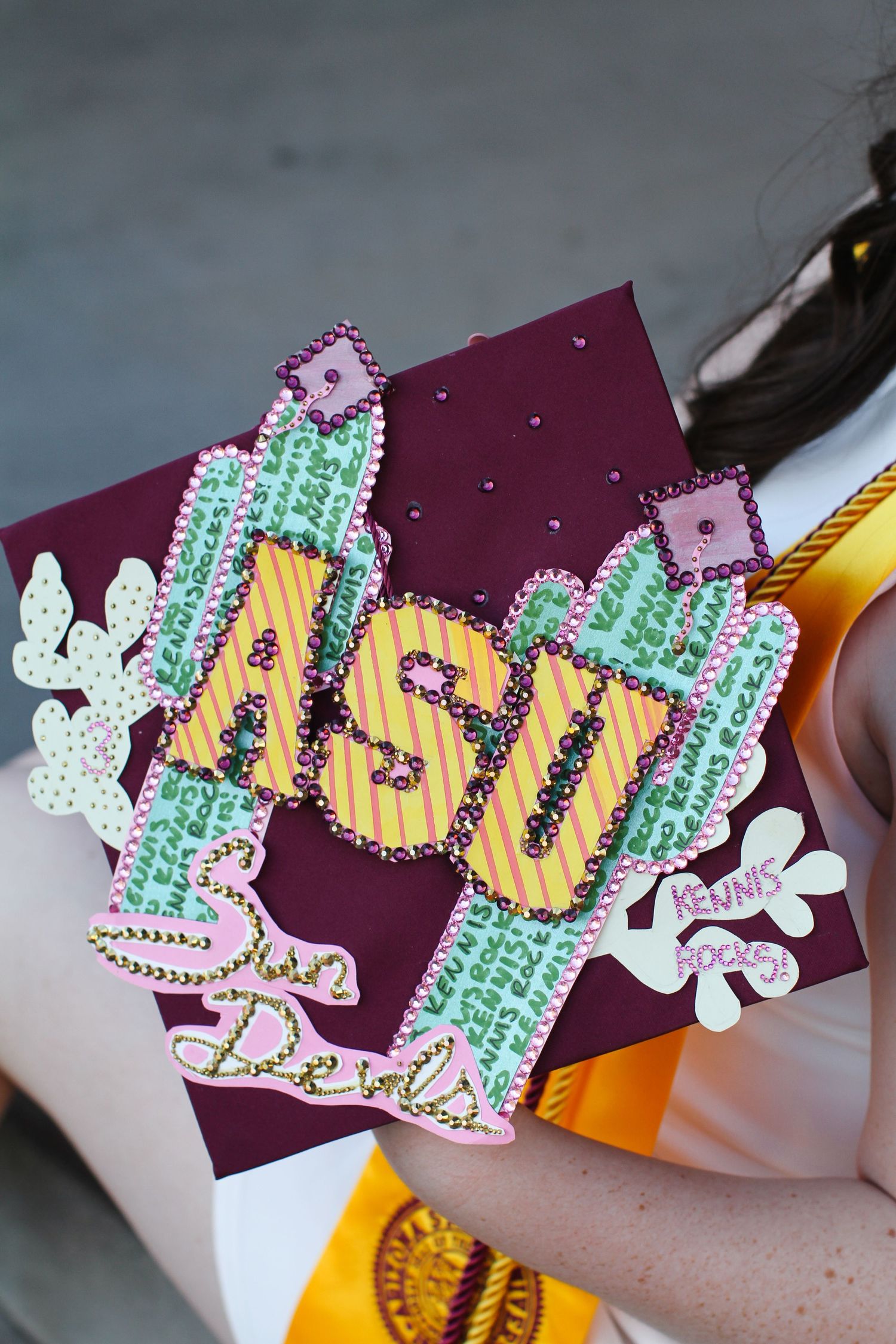 Decorative graduation cap with ASU text design featuring cactus and floral embellishments.