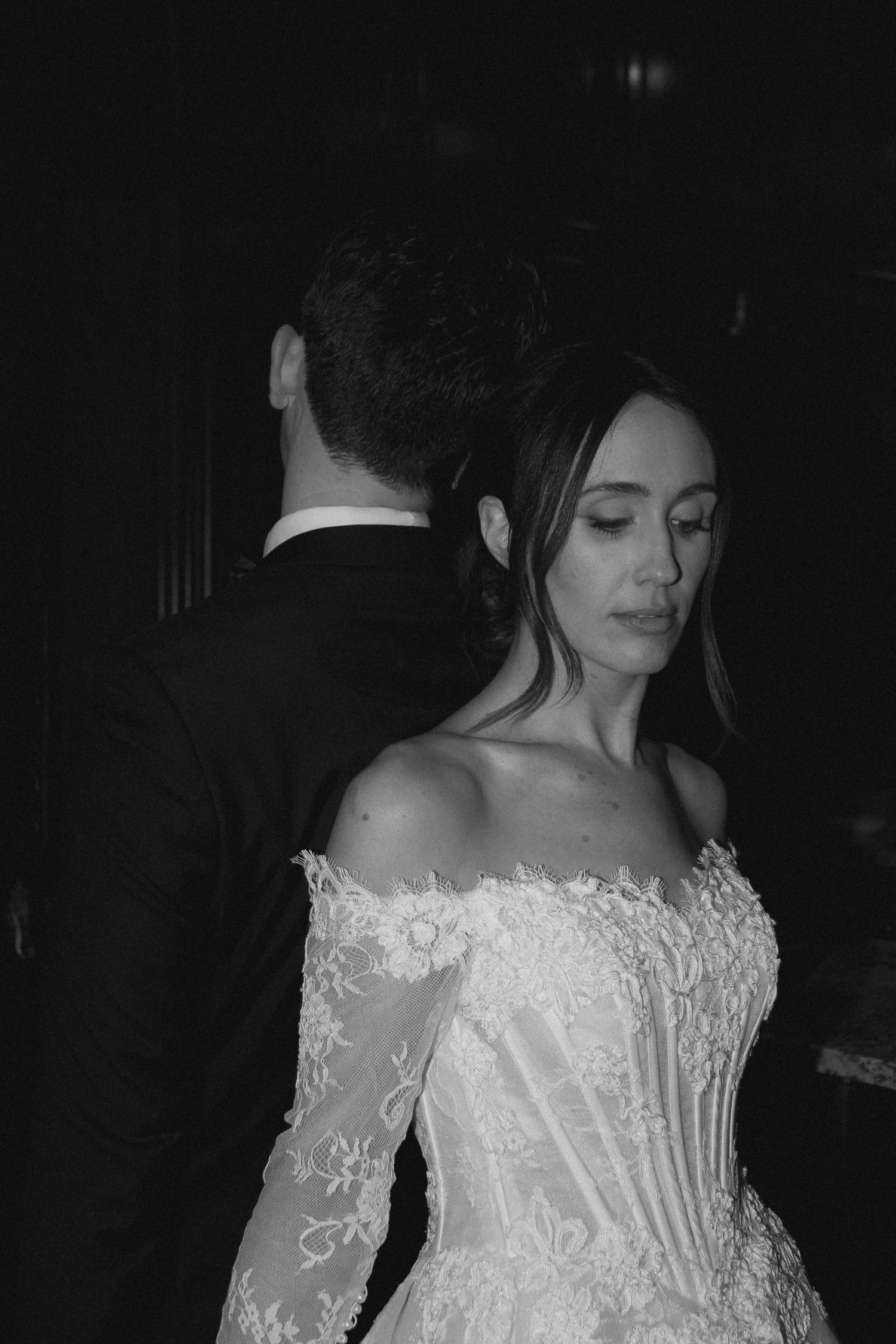 Black and white wedding photo of bride in off-shoulder lace gown standing close to groom in tuxedo.