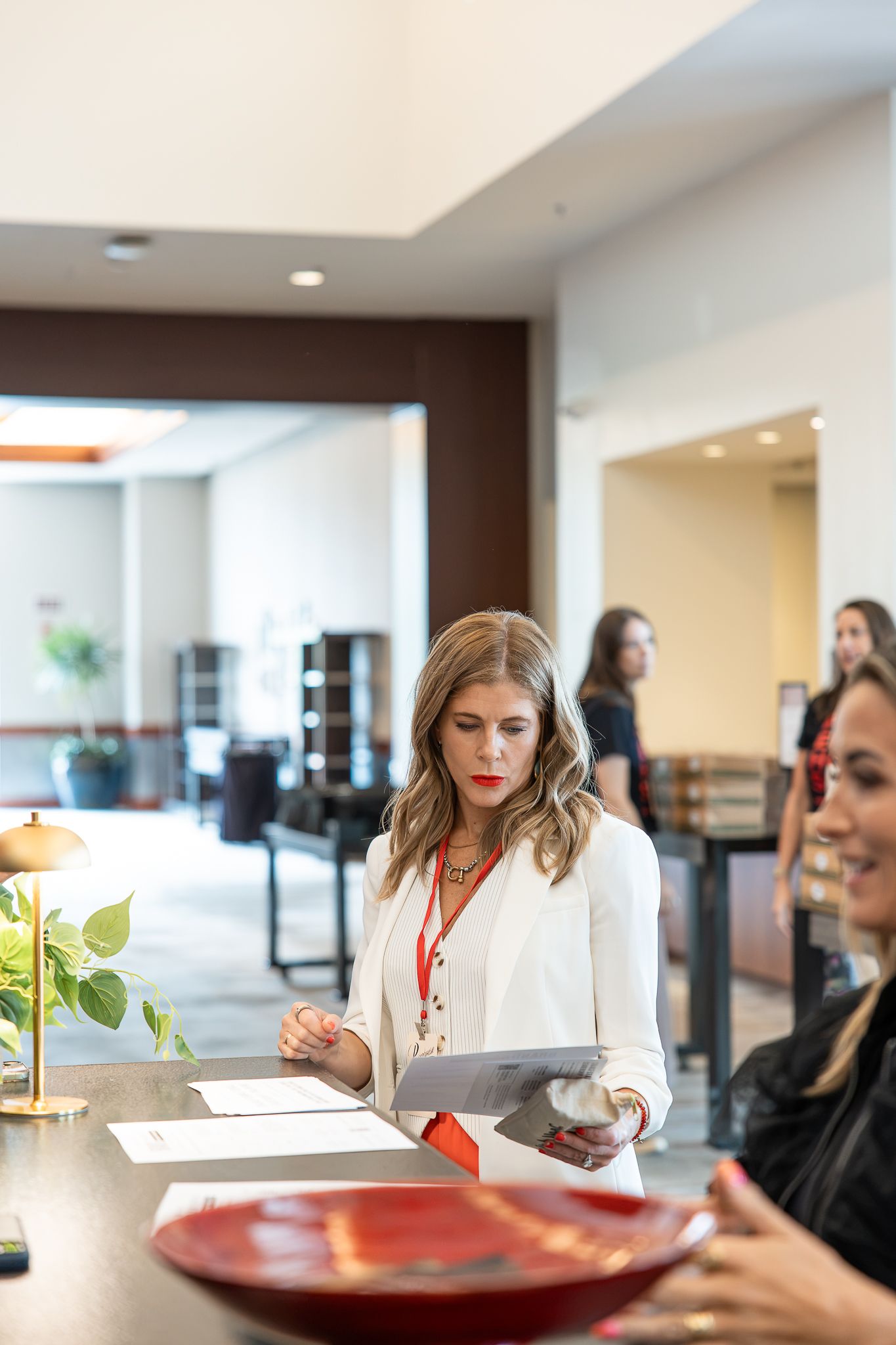 Woman in white blazer reviews documents at a hotel reception desk while colleagues stand in the background.
