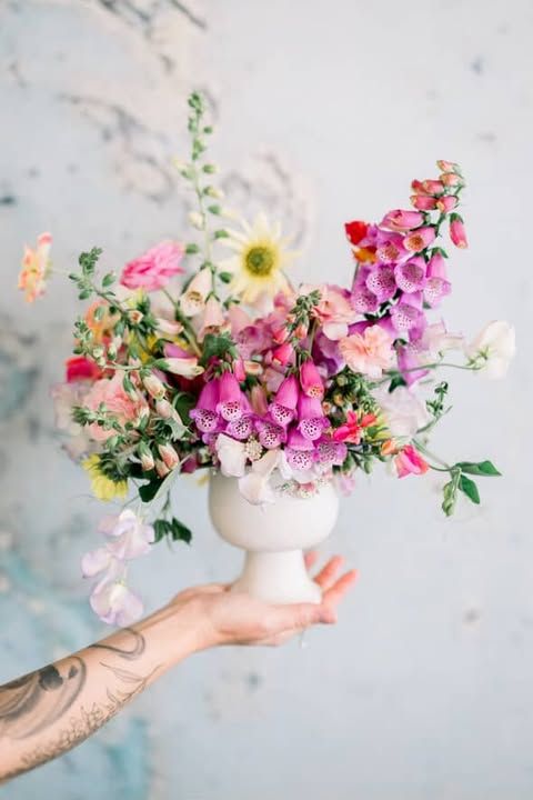 Hands hold a white vase with colorful spring flowers including daisies, peonies and wildflowers against a light blue background.