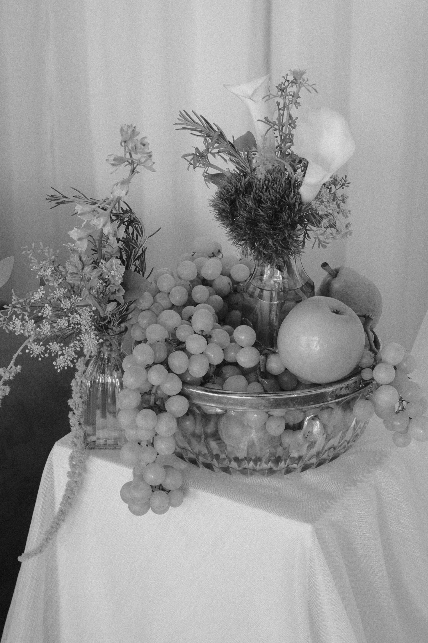 A black and white still life arrangement of grapes, flowers, and vases on a white tablecloth. Tyler Garlej Photography
