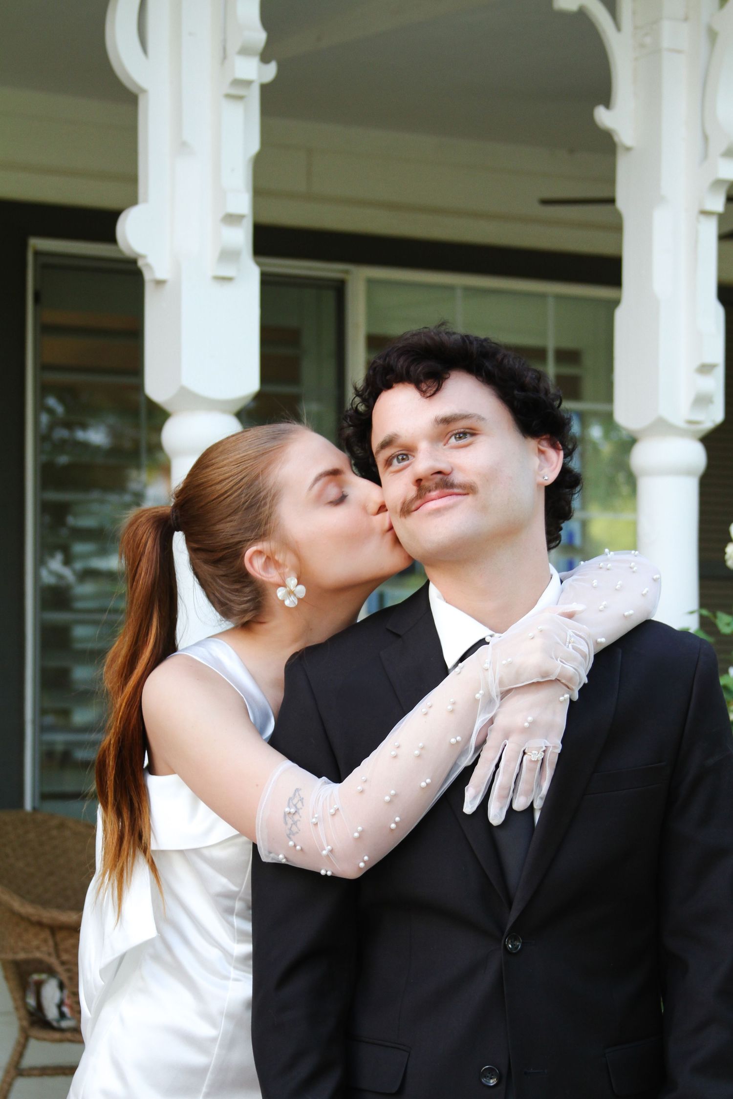 A bride in a white dress and lace gloves kisses a groom in a black suit on the cheek outdoors.