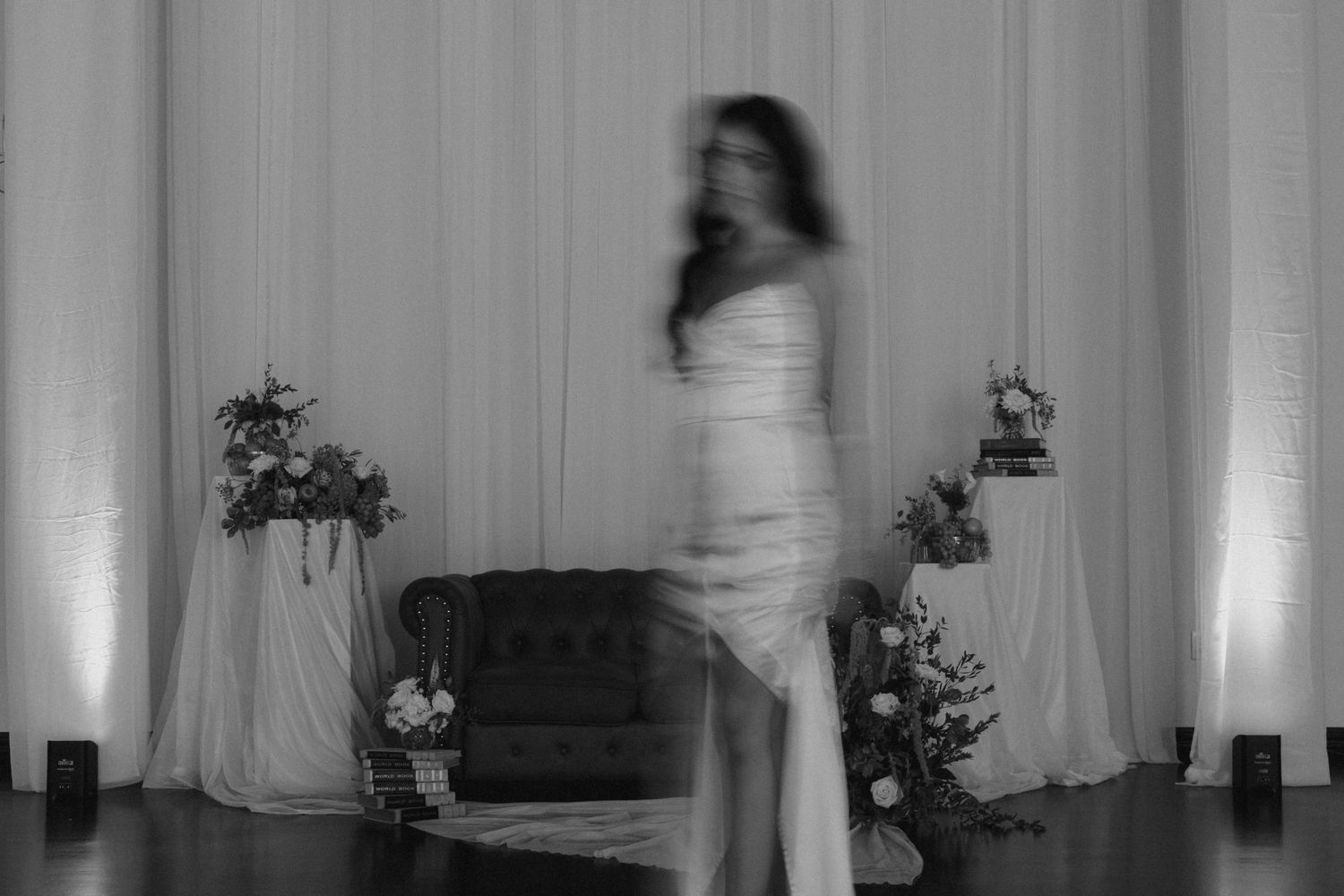 Blurred black and white photo of a woman in a wedding gown posing elegantly among floral arrangements.