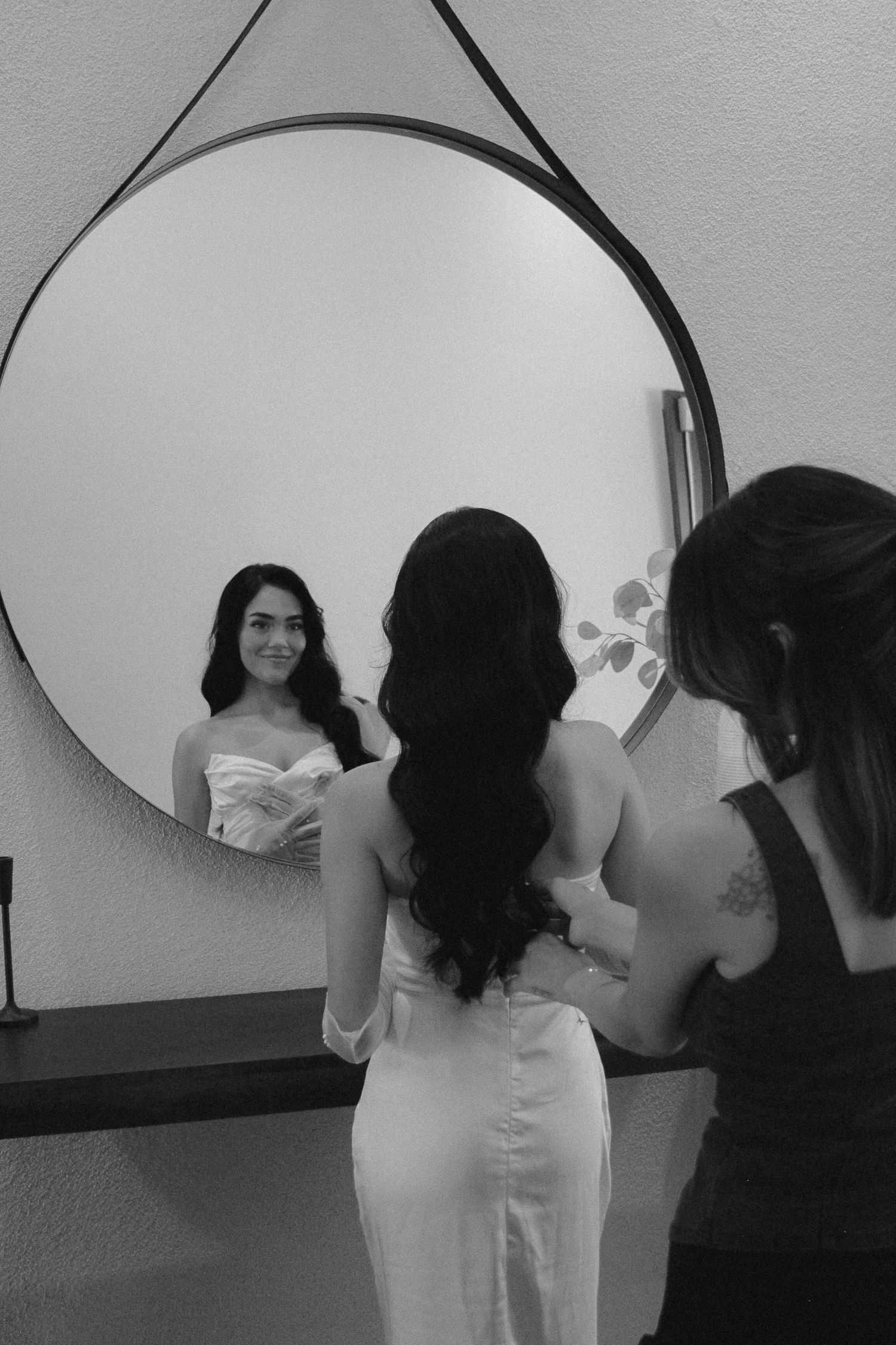 Bride in white gown smiles at her reflection in a round mirror while bridesmaid helps her dress.