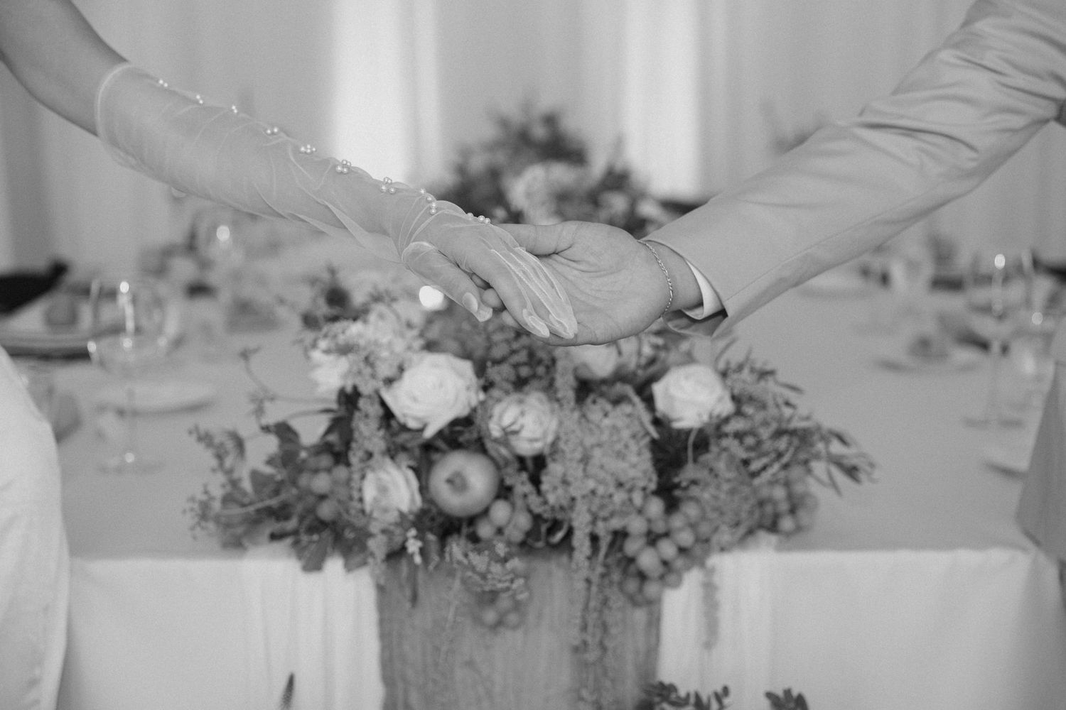 Black and white photo of bride and groom holding hands over floral centerpiece at wedding reception table.