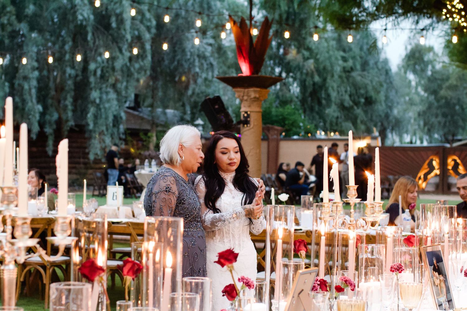 An elegant outdoor wedding reception setup with string lights, candles and red floral arrangements in a garden setting.