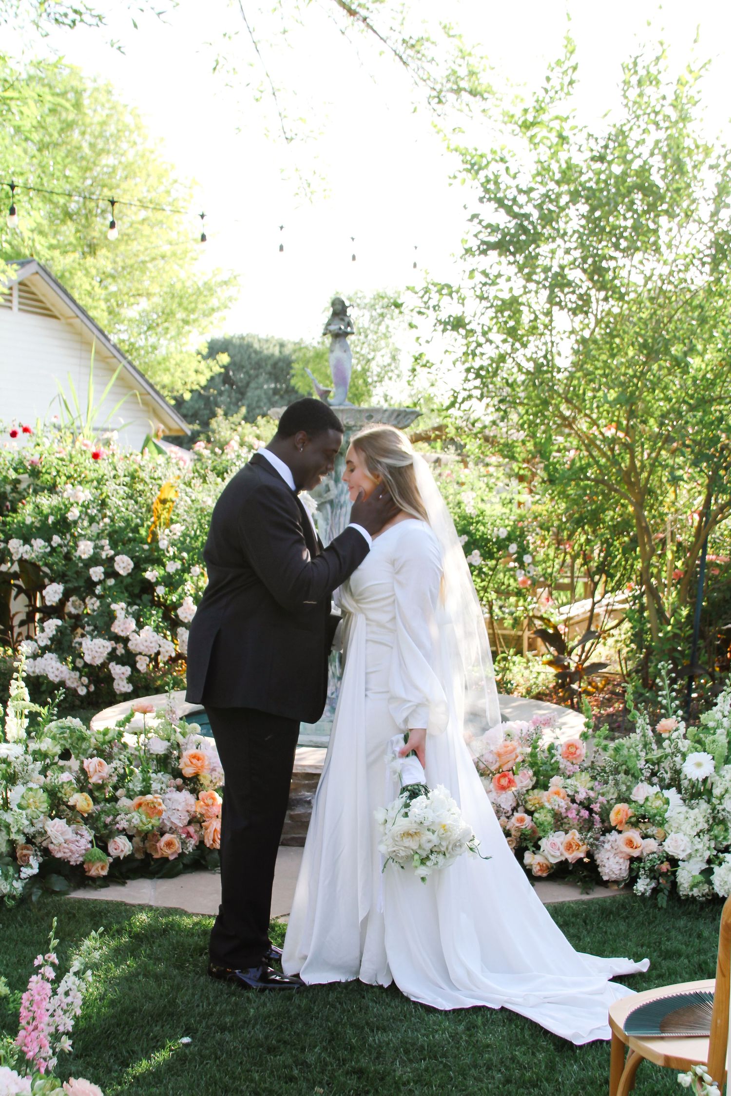 A wedding couple shares an intimate moment in a garden surrounded by beautiful pink and white roses and greenery.