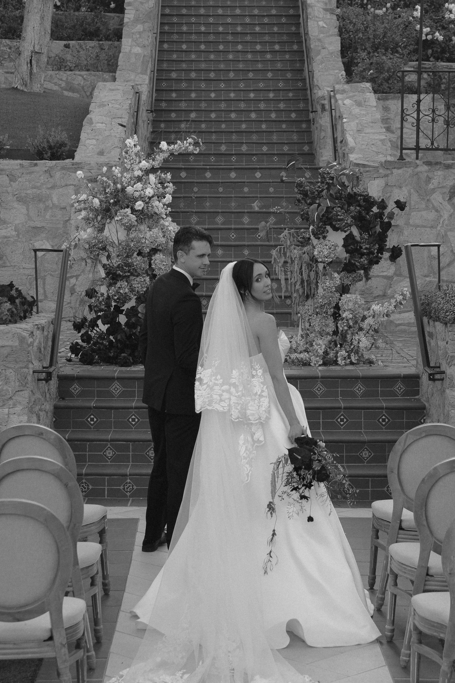 Black and white wedding photo of bride in veil and groom in suit standing before floral-adorned staircase.
