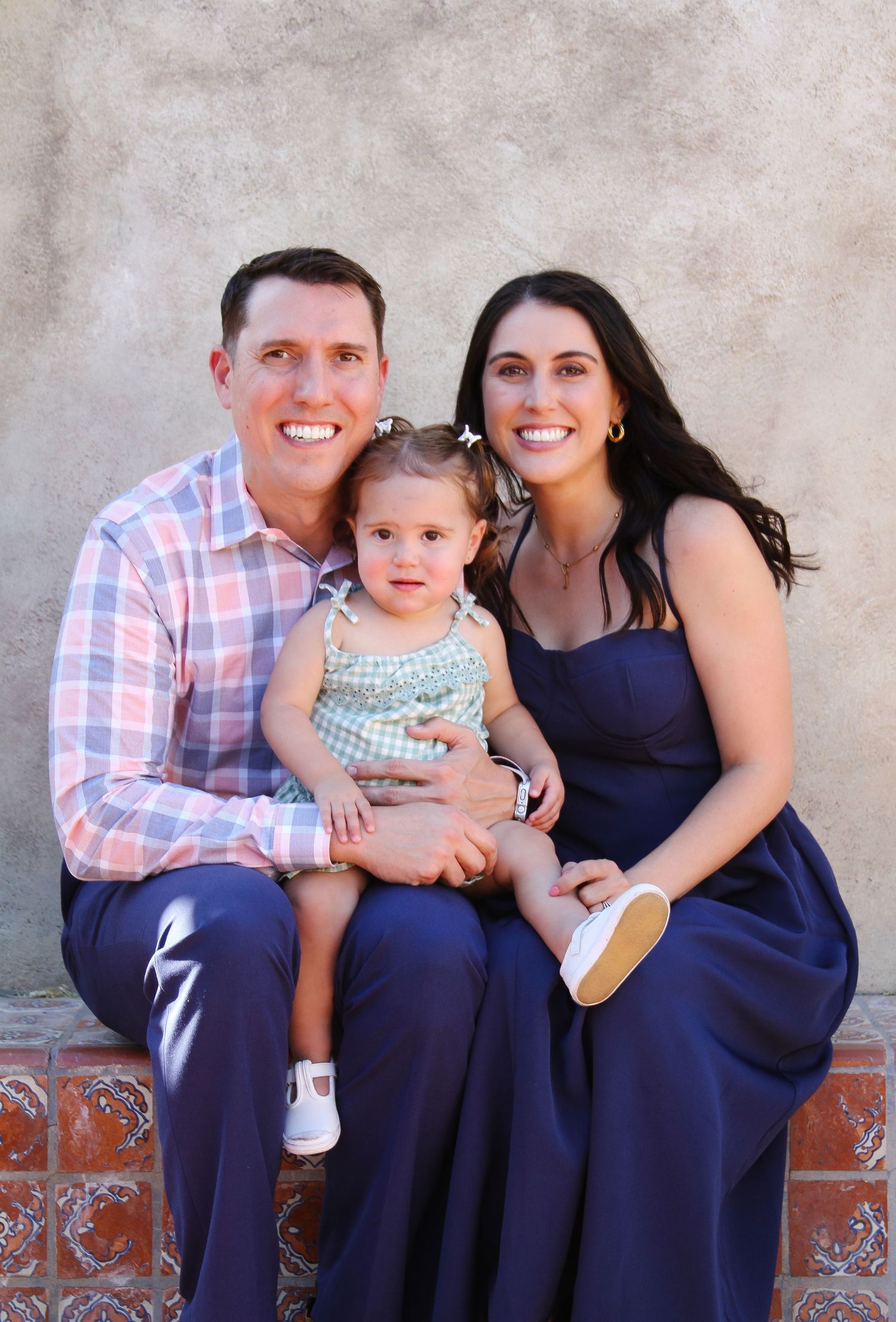 Family portrait with young child in blue and pink outfits against neutral backdrop.