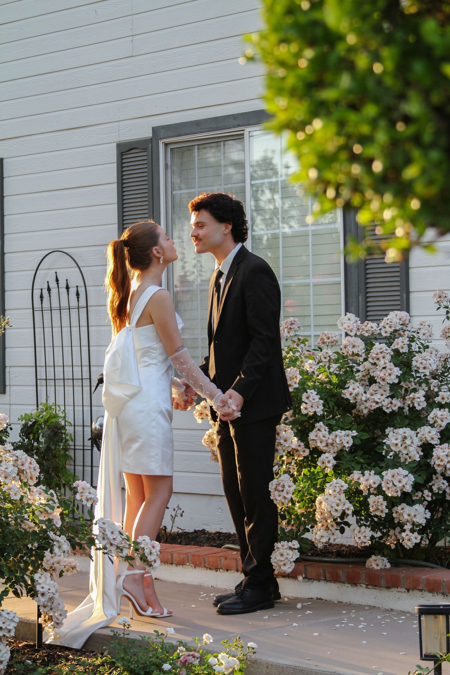 Bride and groom holding hands outdoors near white house with blooming roses, sharing a romantic moment in wedding attire.