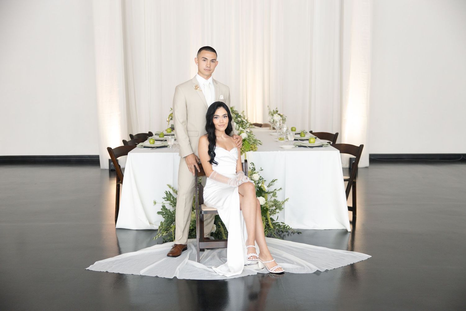 Elegant bride and groom pose before a white draped sweetheart table with lush greenery centerpieces.