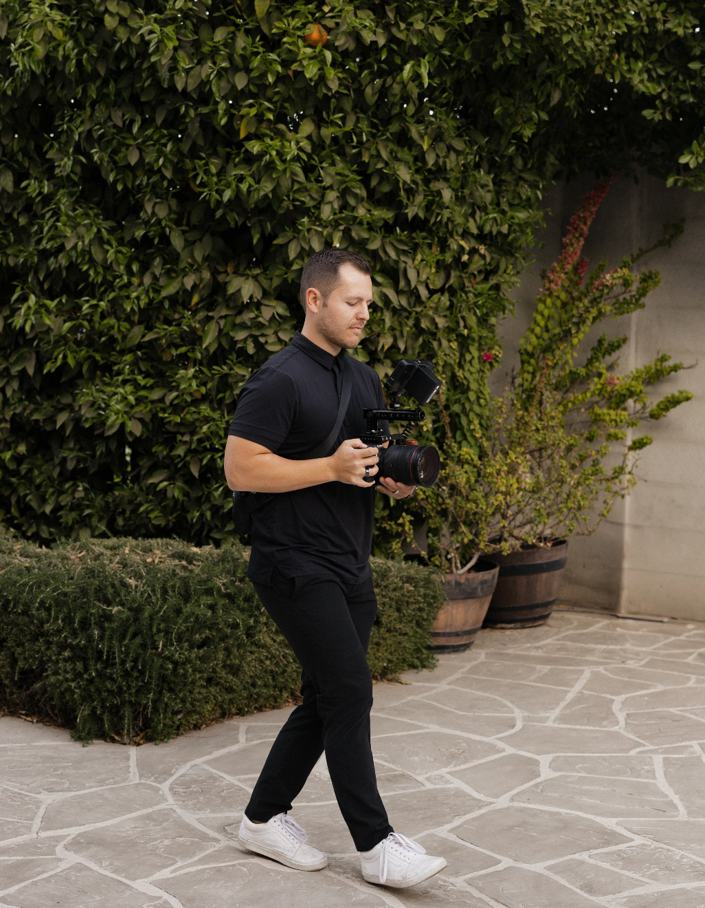 Photographer in all black attire walking with camera gear near garden setting.