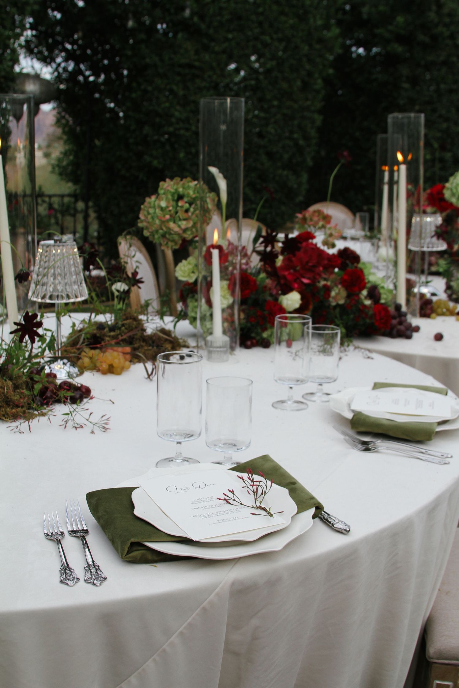 Elegant outdoor wedding reception table setting with red roses, green napkins, crystal glasses, and candlelight centerpieces.
