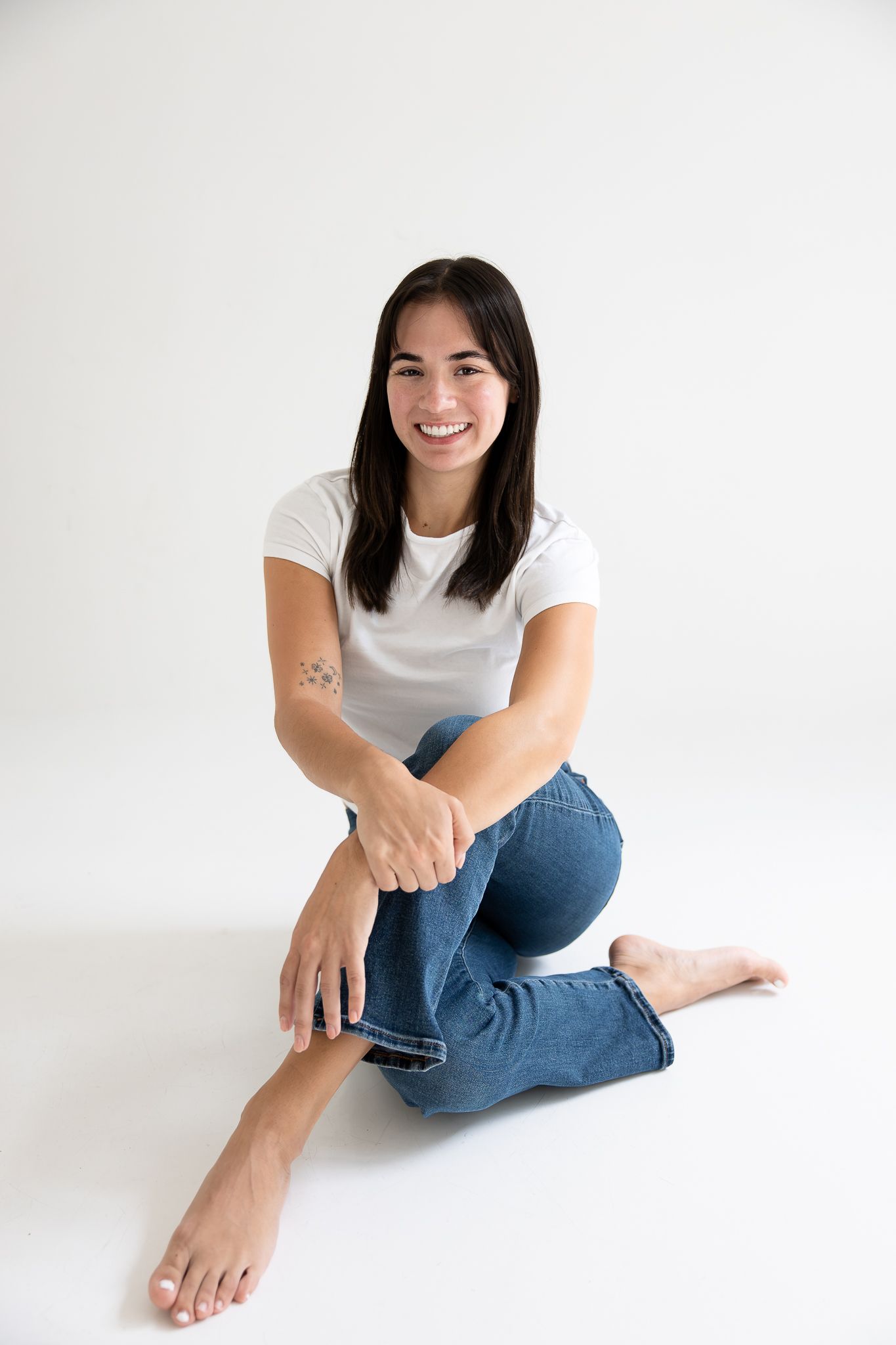 Smiling young woman sitting on white floor in white t-shirt and blue jeans, barefoot with a small tattoo on her arm.