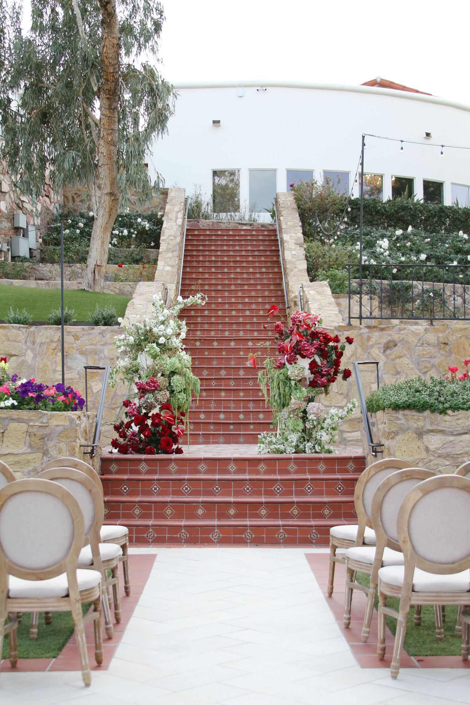 Outdoor wedding ceremony aisle with red tiled stairs adorned with red and white floral arrangements.