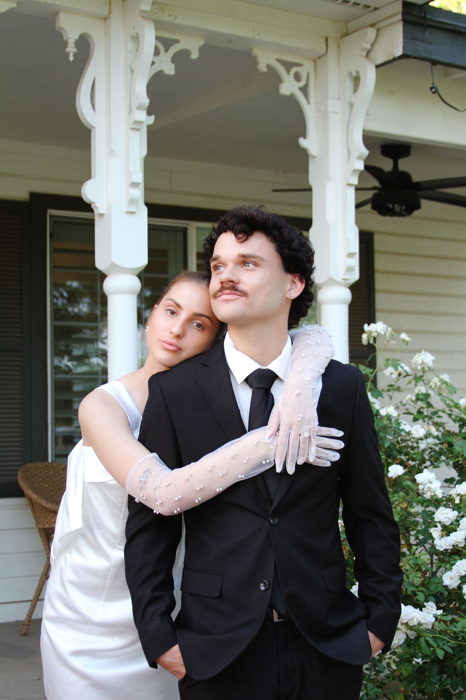A couple in wedding attire poses before a white Victorian porch, bride in gloves leaning on groom in black suit.