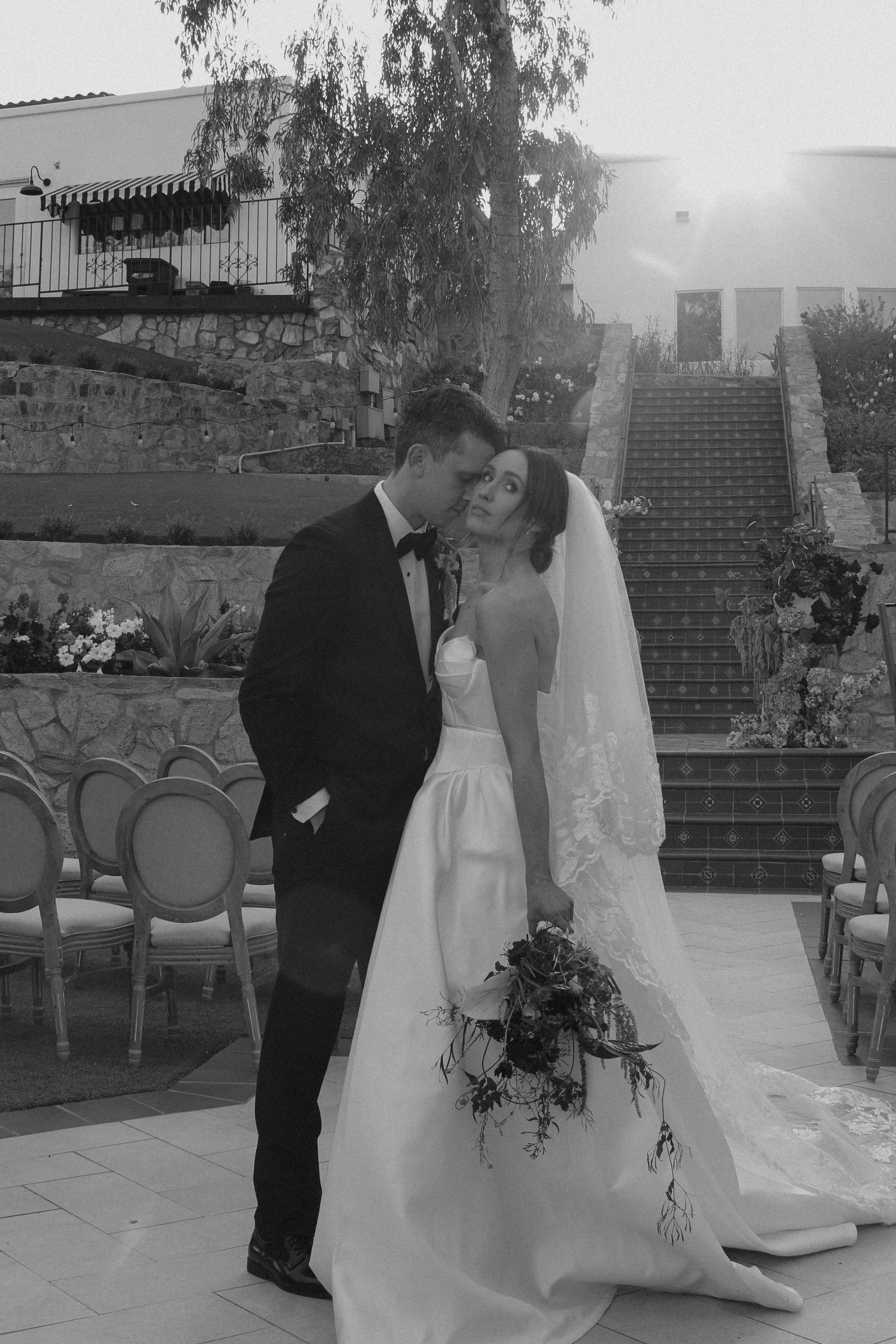 Black and white photo of a bride and groom sharing a kiss at an outdoor wedding ceremony with stone steps.