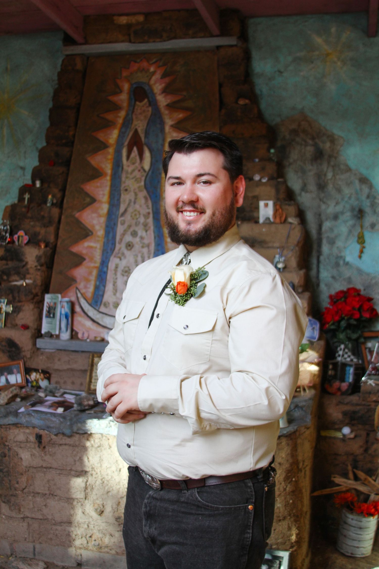 Groom in cream shirt smiles in front of decorative mural with religious artwork.