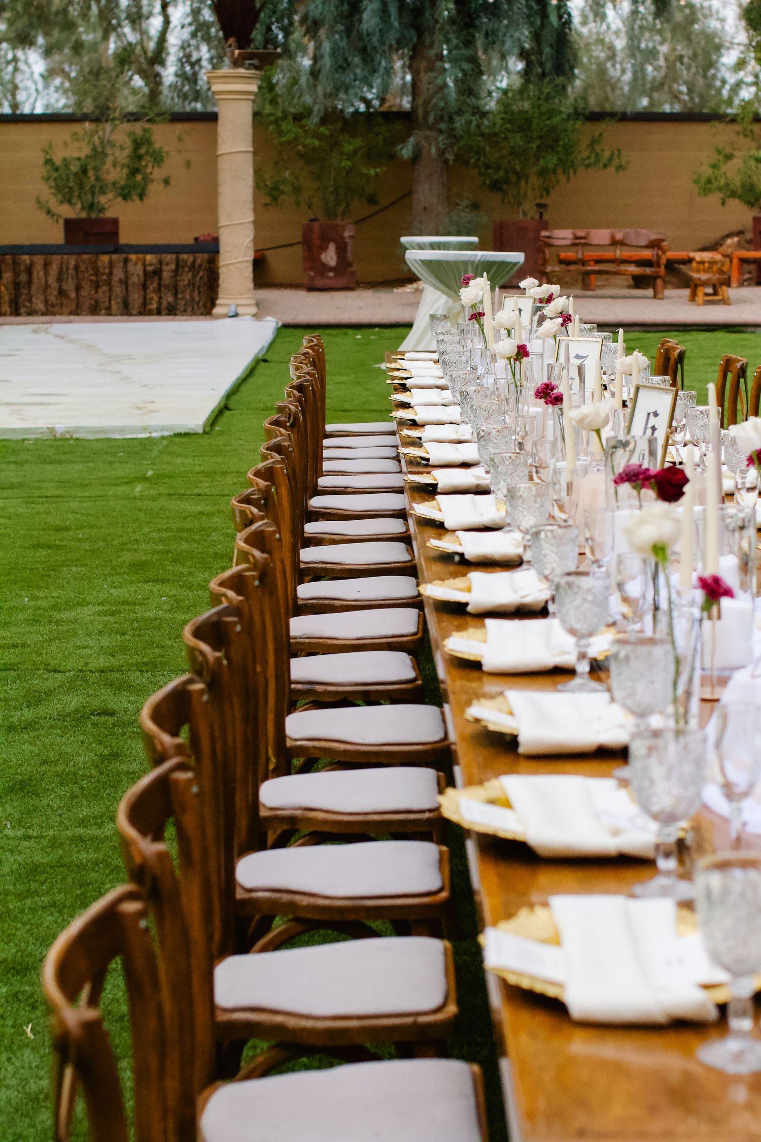 Elegant outdoor dining setup with long wooden table featuring white cushioned chairs and delicate floral centerpieces.