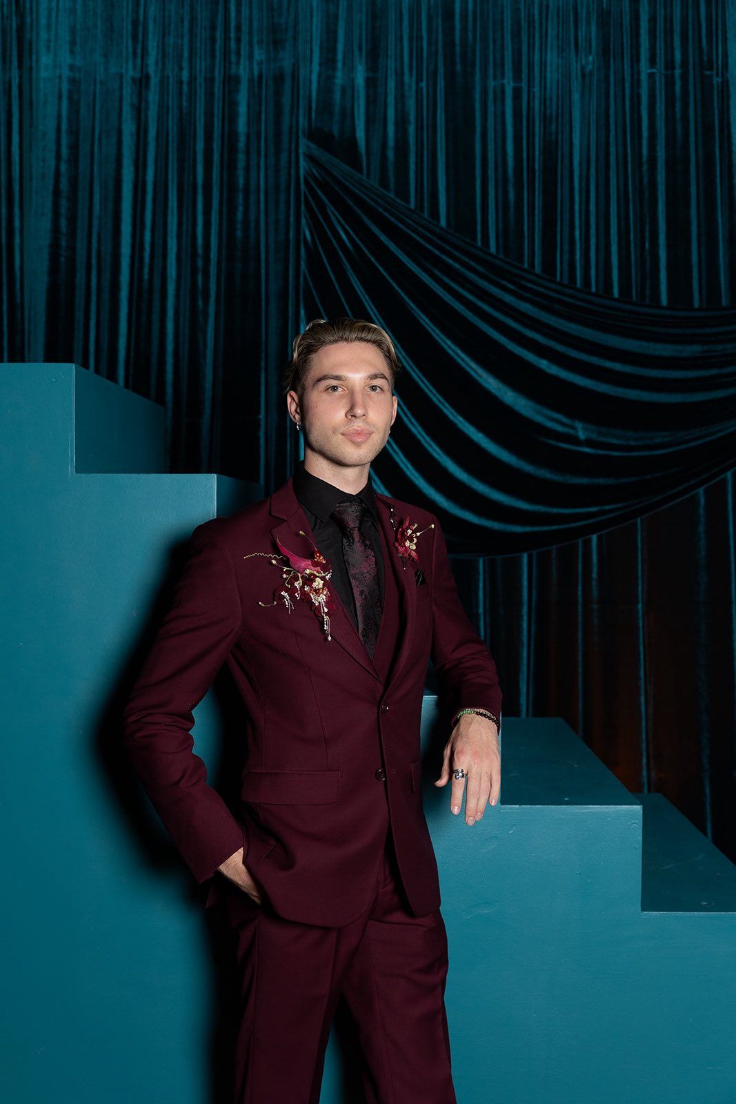 Young man in burgundy suit with floral embroidery poses on teal stairs against dark velvet curtain backdrop.