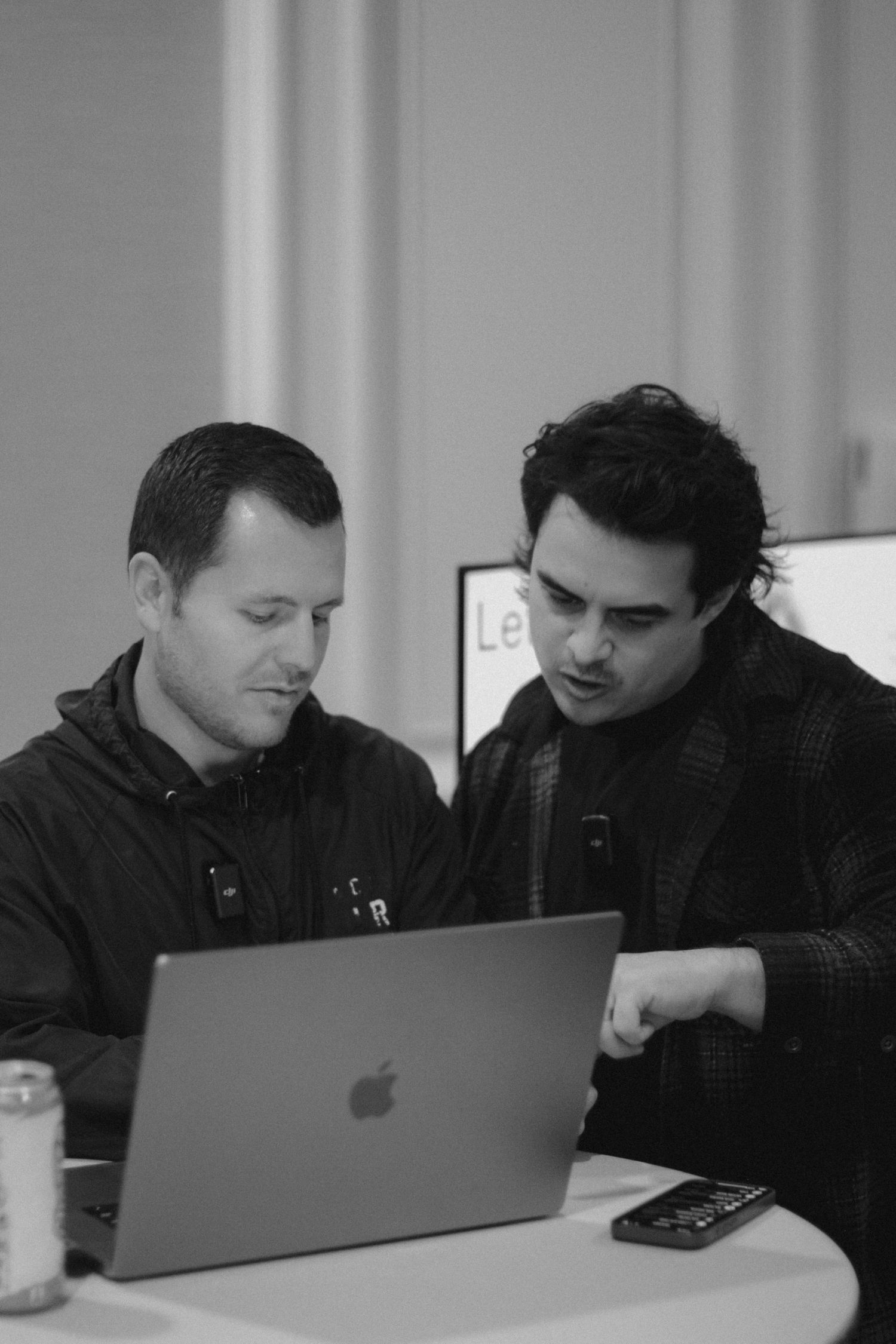 Two men collaborating over a MacBook laptop in a black and white office setting, one pointing at the screen.