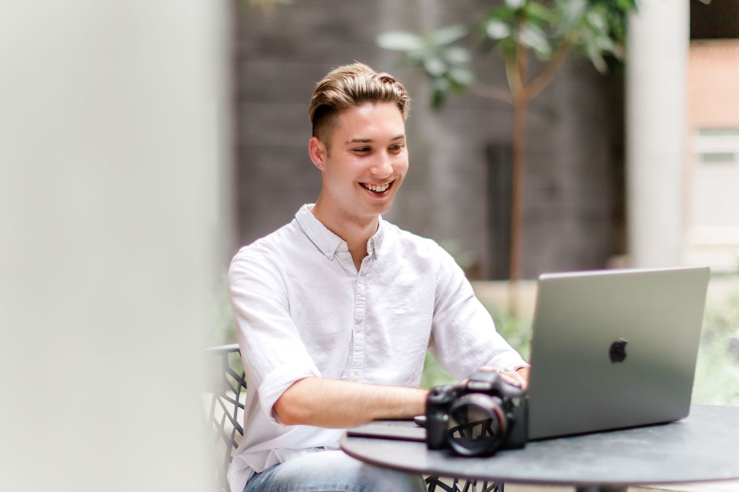 Photographer in white shirt working outdoors on laptop with camera on table.