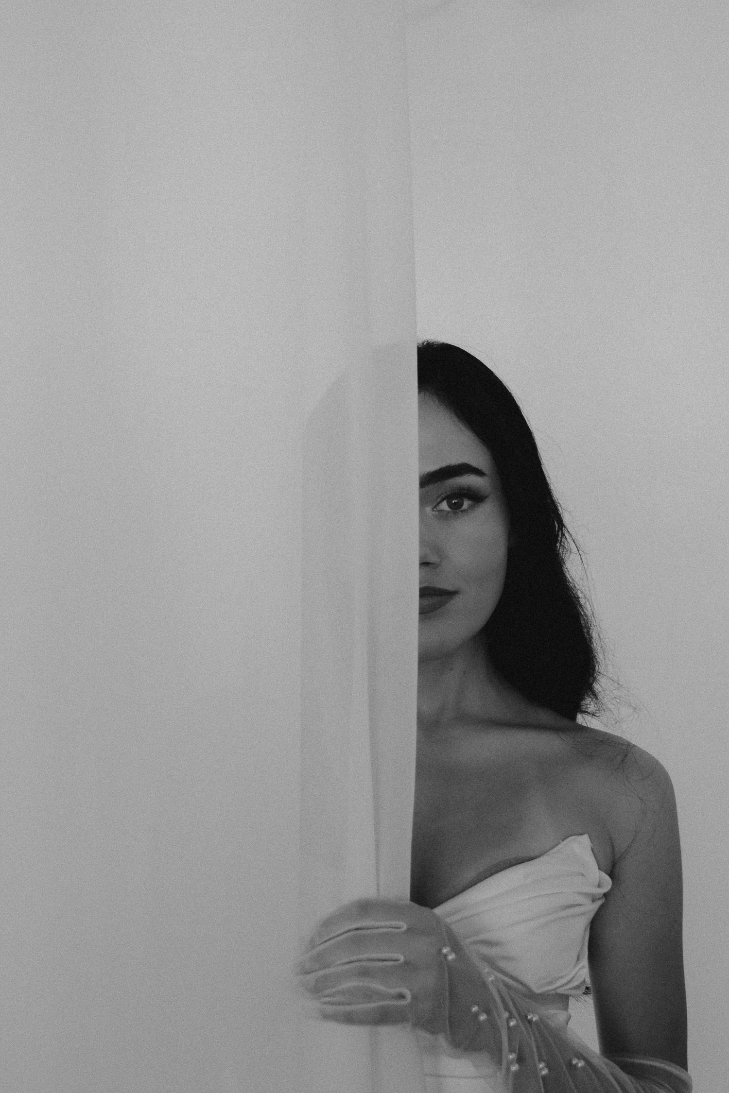 Elegant bride in a strapless gown peeks around a white door in a classic black and white wedding portrait.