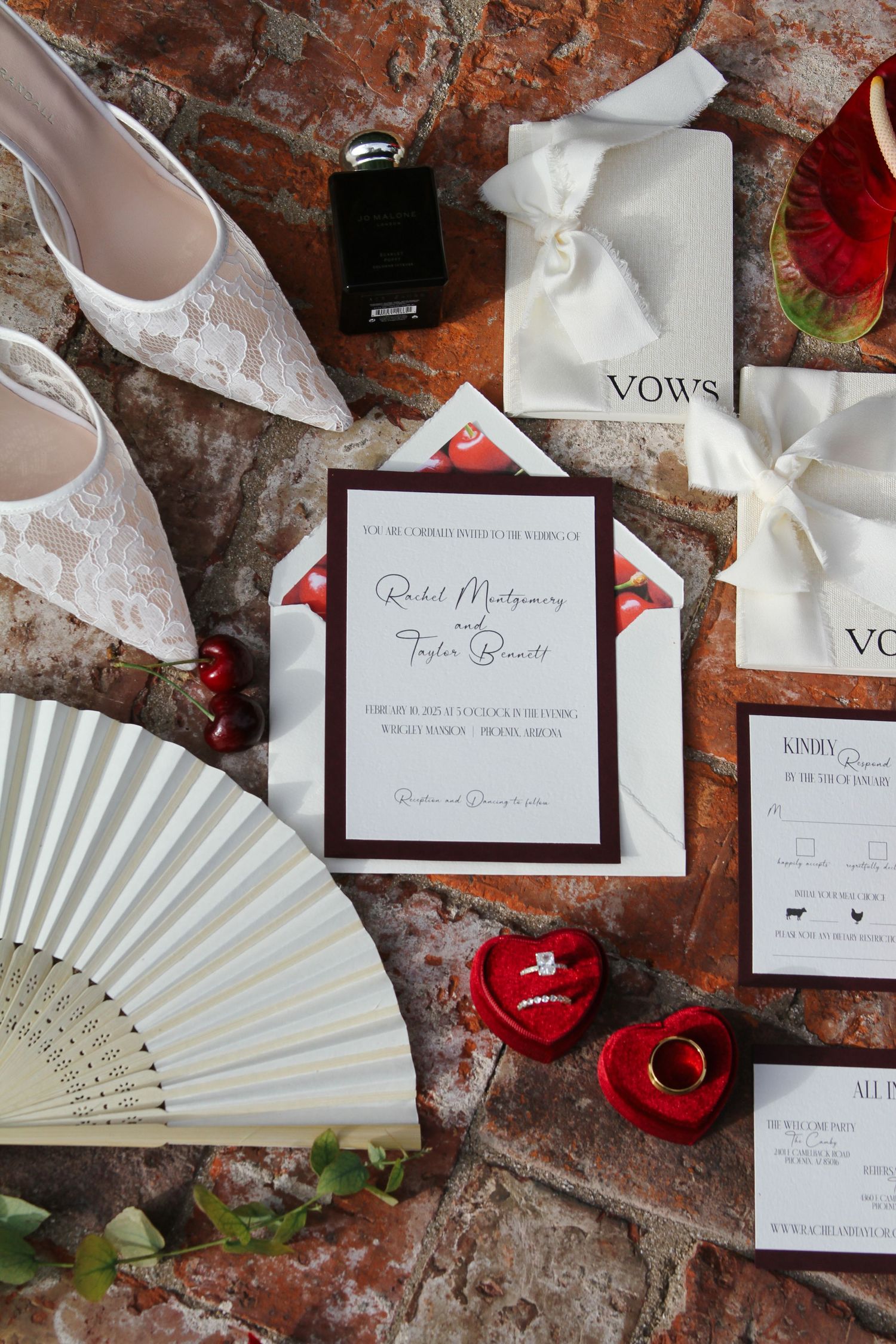 Bridal flat lay with lace heels, invitation suite, vow books, red ring boxes, and white fan on brick.