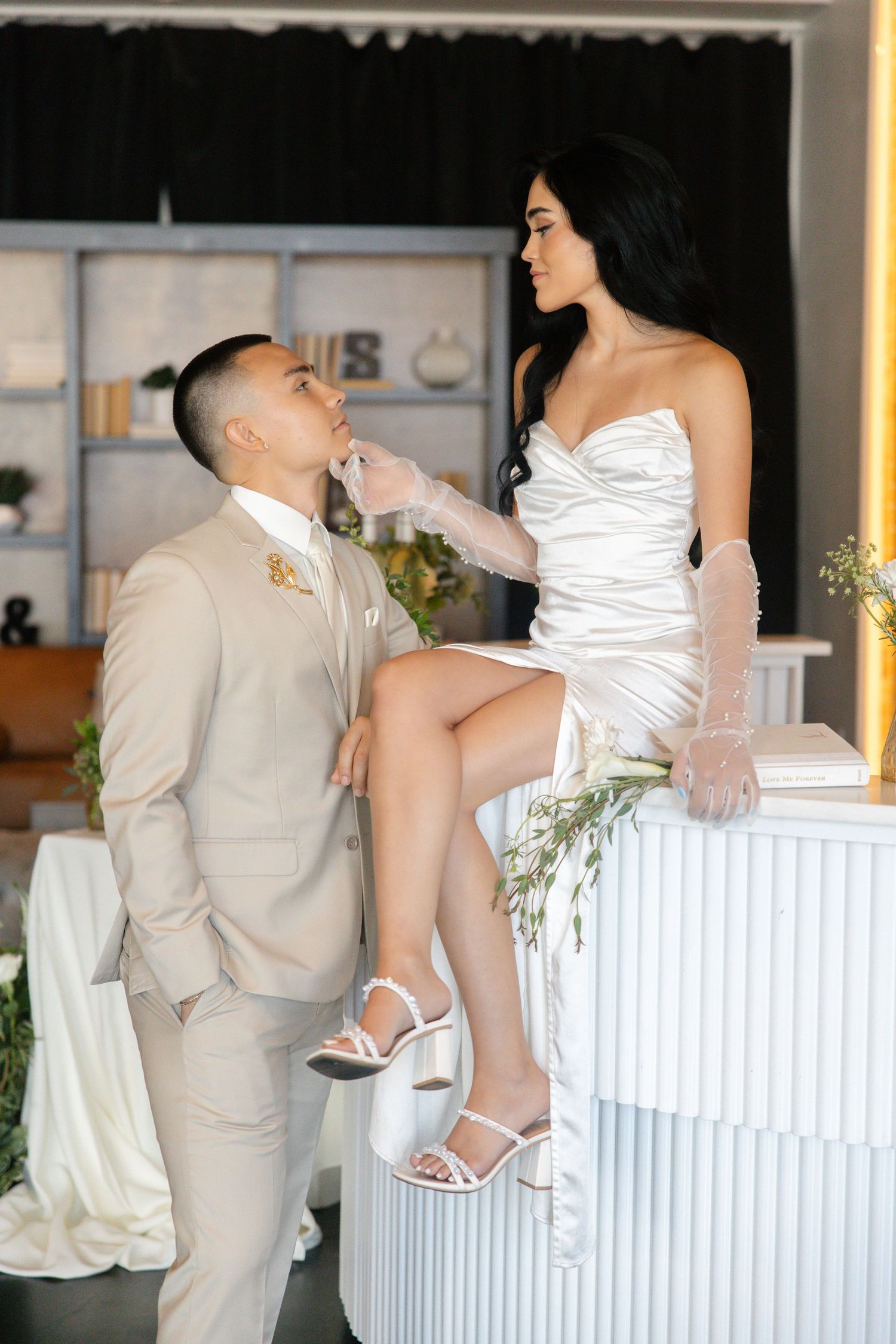 Bride in white strapless dress sits on pedestal as groom in beige suit kisses her hand at wedding.
