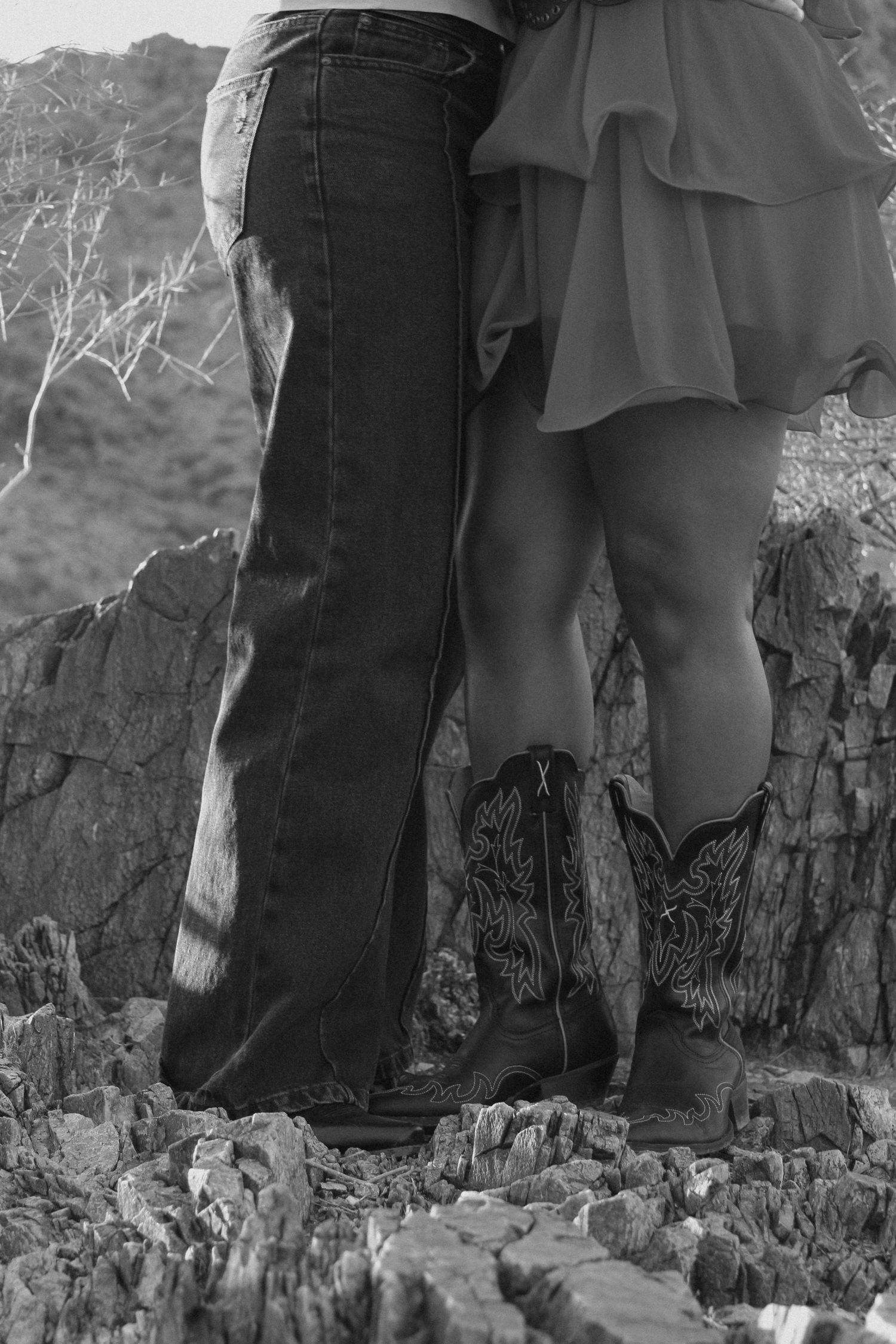 A couple wearing contrasting outfits and cowboy boots stand together on rocky terrain.