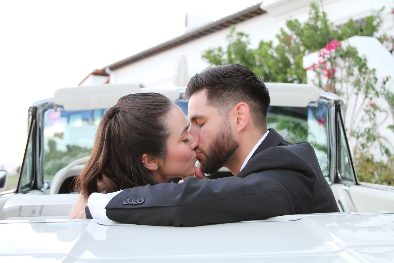 Newlyweds share a romantic kiss while sitting in a vintage white car.
