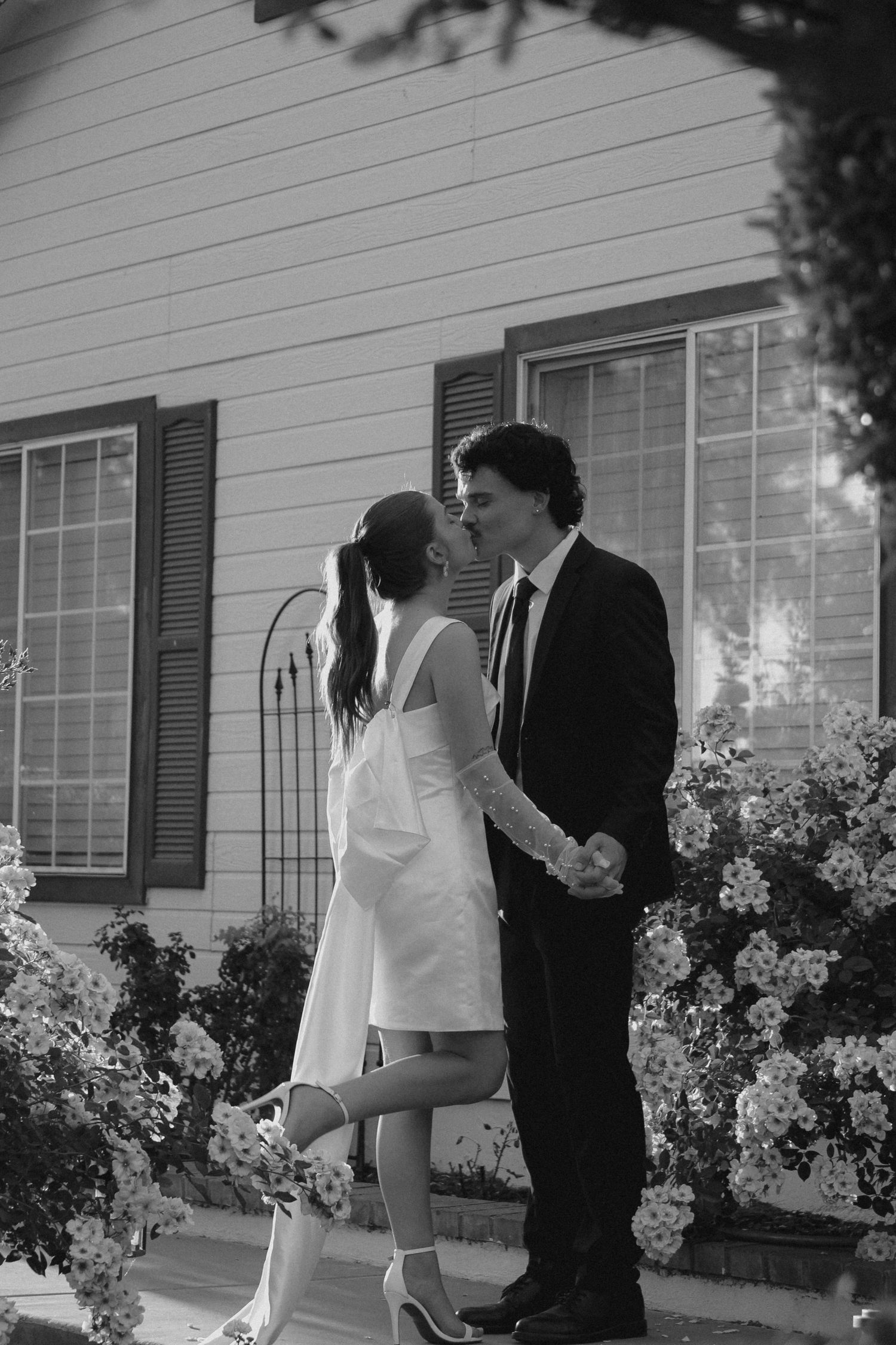 Black and white photo of a couple kissing in formal attire surrounded by rose bushes outside a classic home.