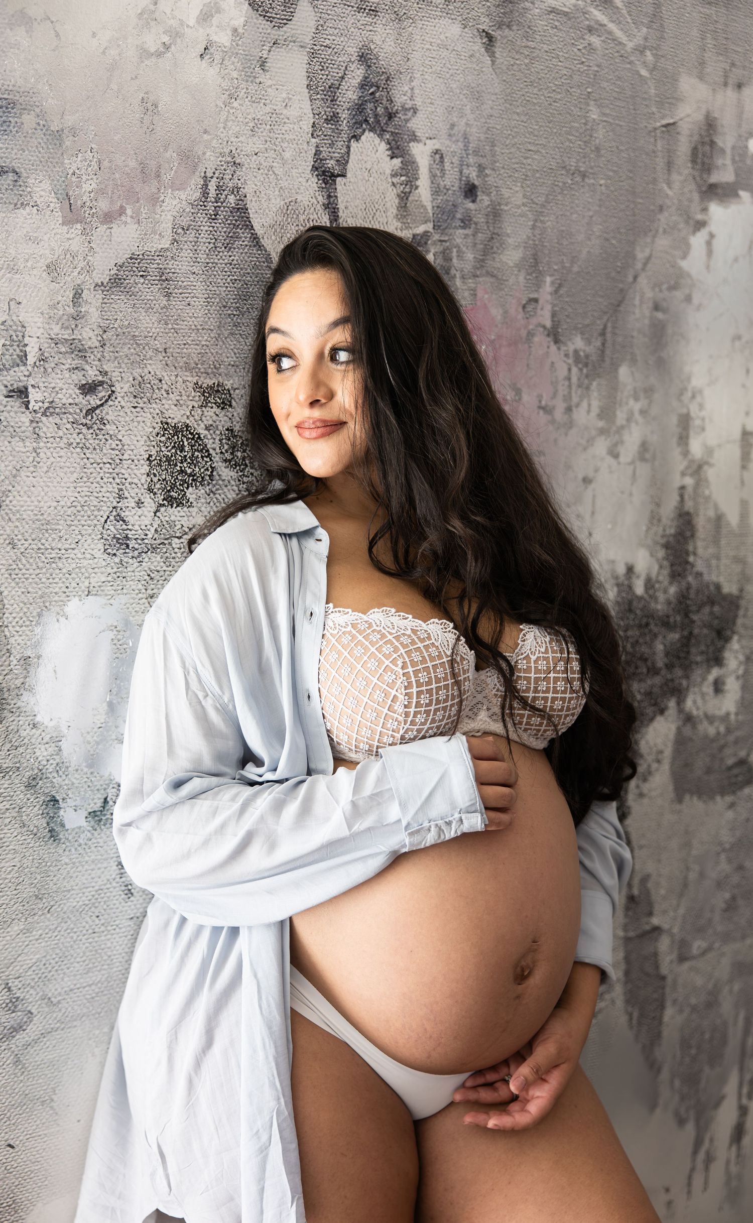 Pregnant woman in white lace bralette and open blue shirt cradling baby bump against abstract gray wall.