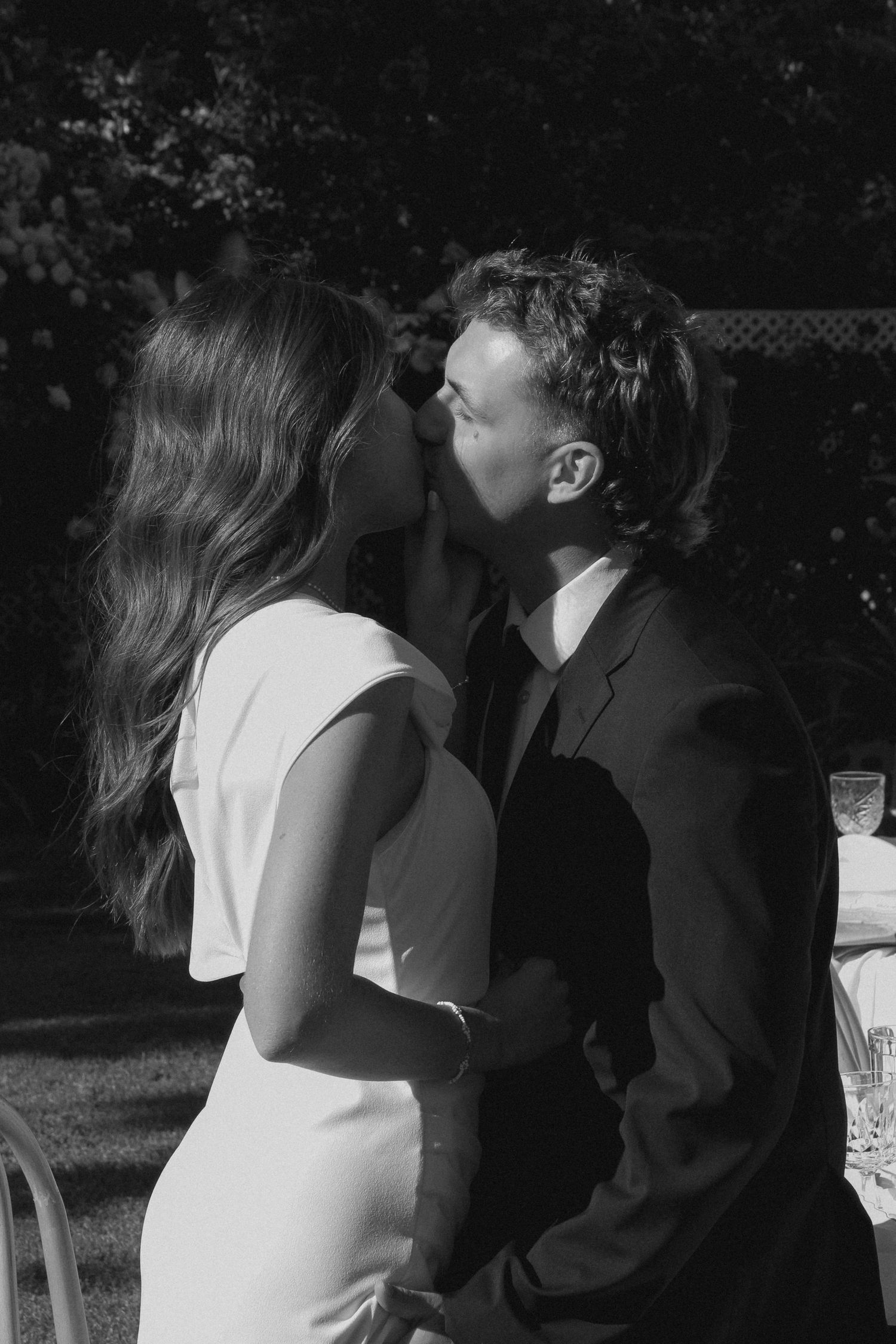 A black and white photograph capturing an intimate wedding moment in a garden setting.