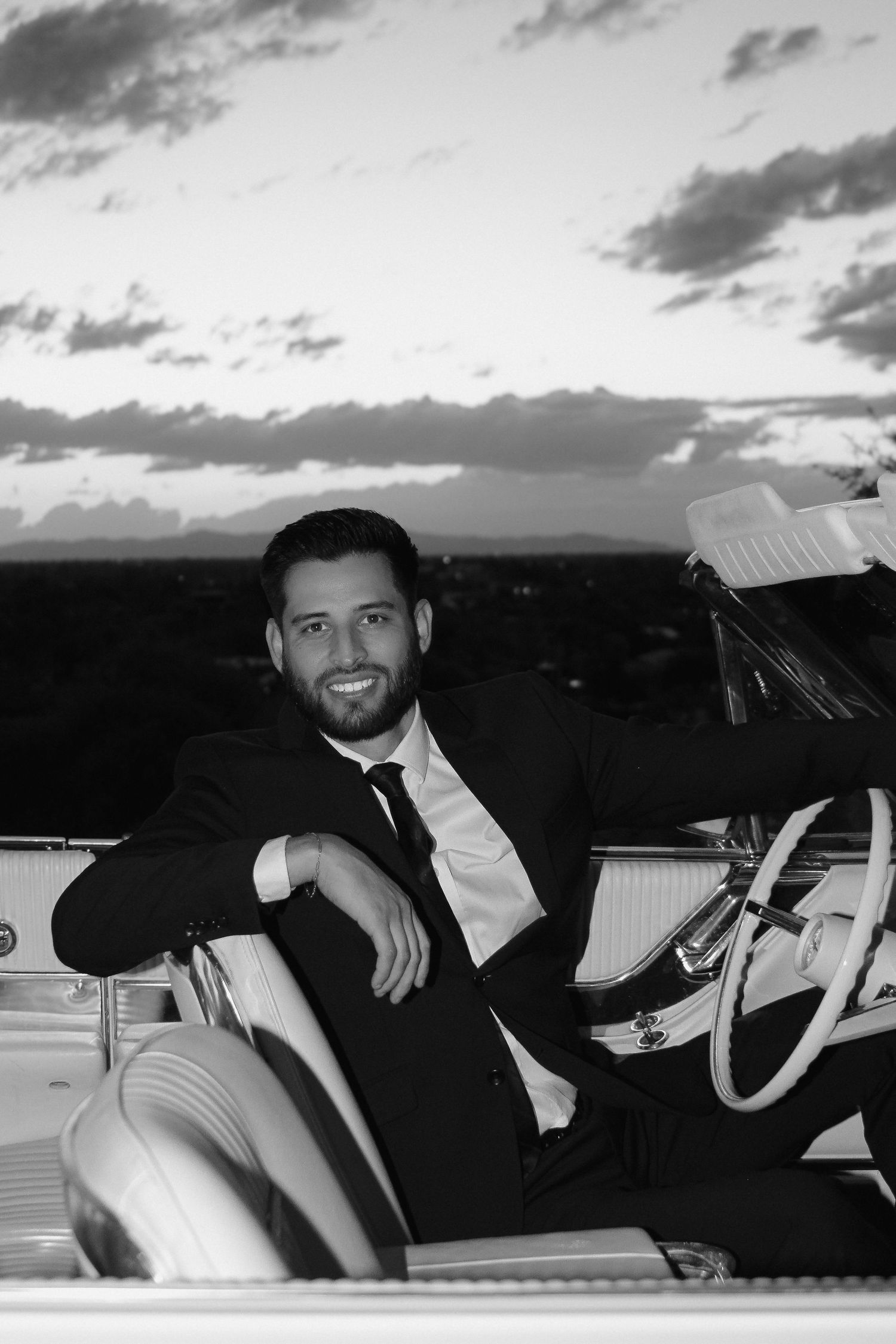 Black and white portrait of groom in suit sitting in vintage car at sunset.