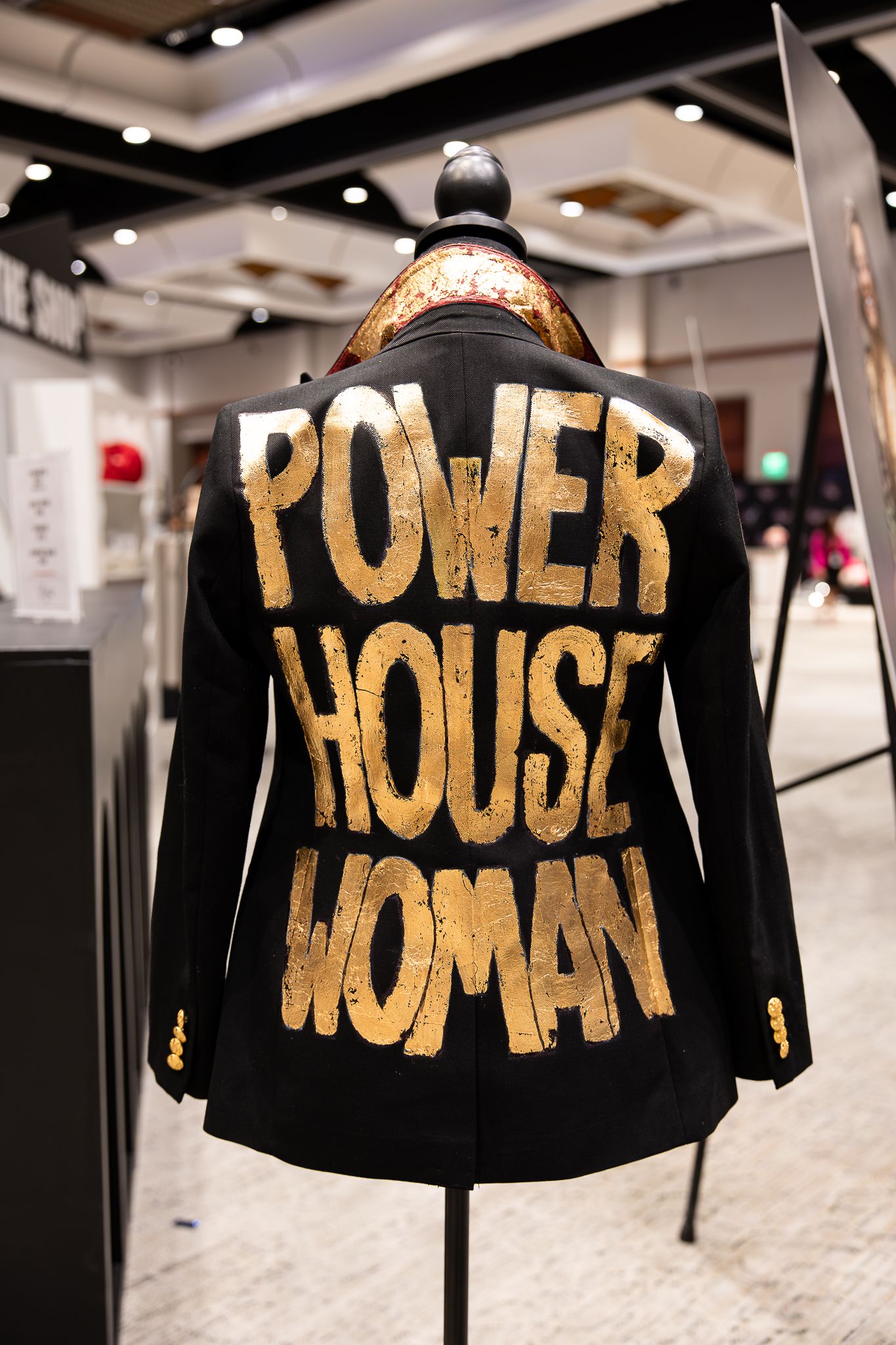Black blazer on mannequin with gold painted 'Power House Woman' text displayed at fashion exhibition.