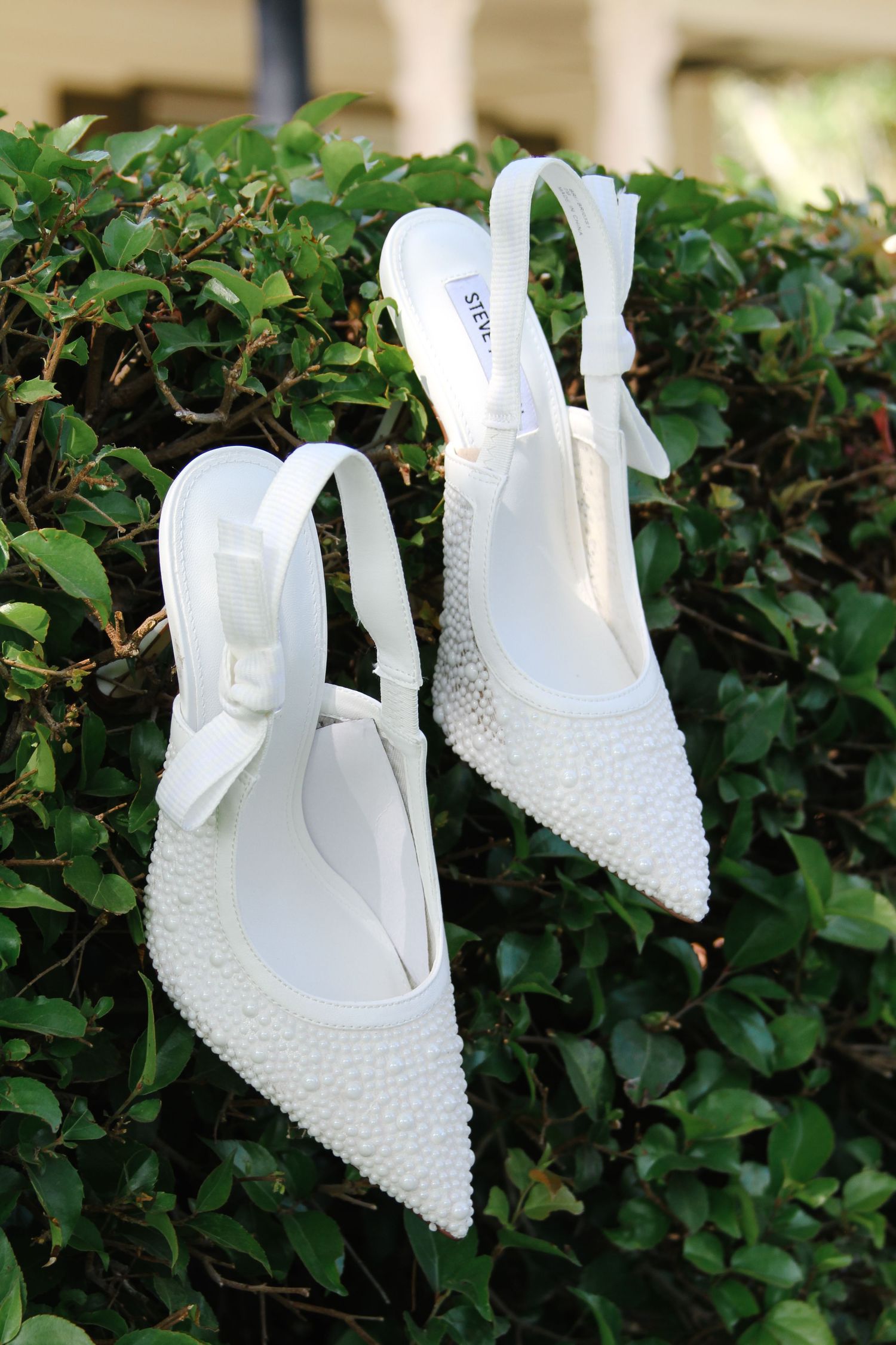 A pair of elegant white beaded bridal heels displayed outdoors against lush green foliage in natural lighting.