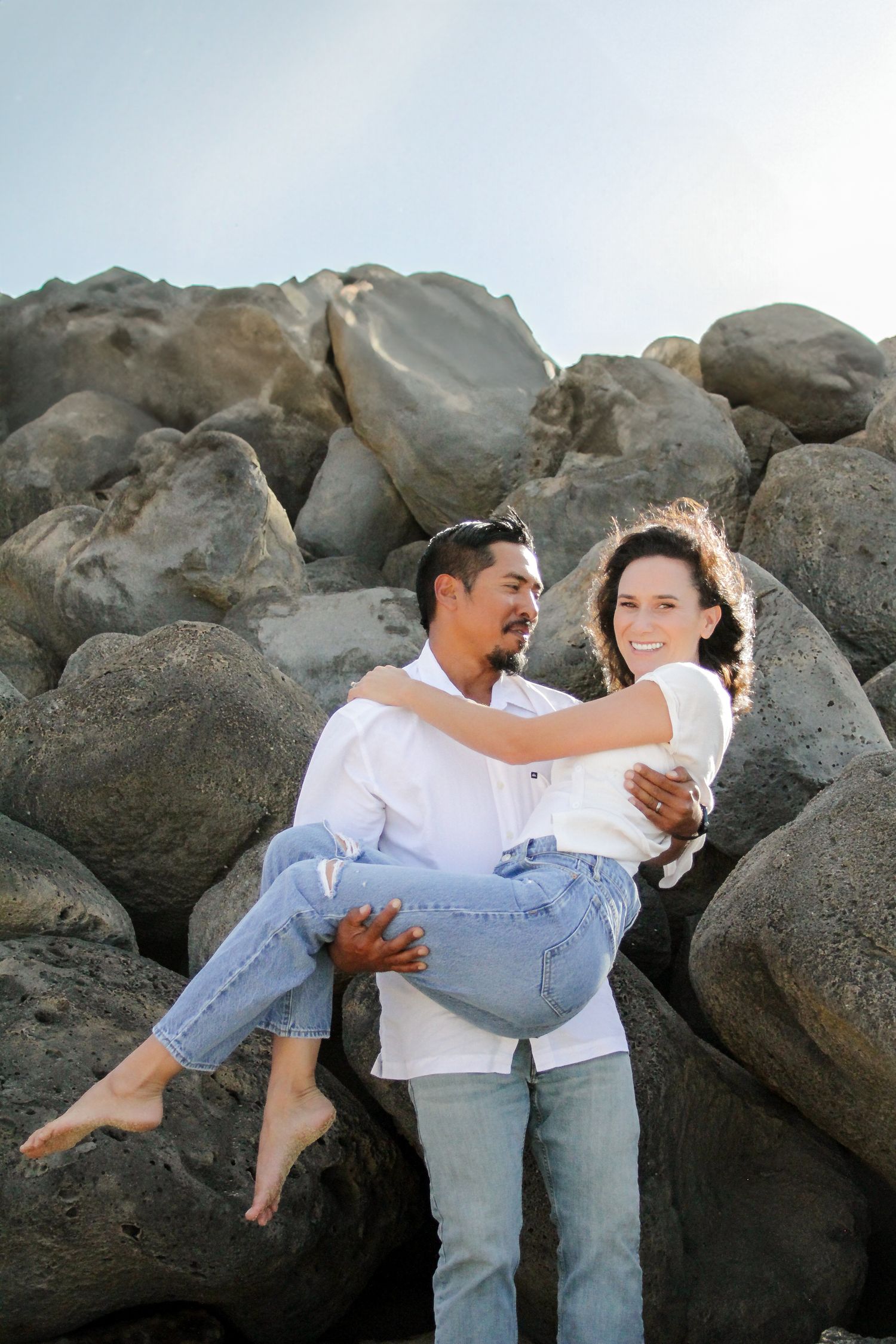 A couple embraces and smiles together while posing on rocky coastal terrain at sunset.