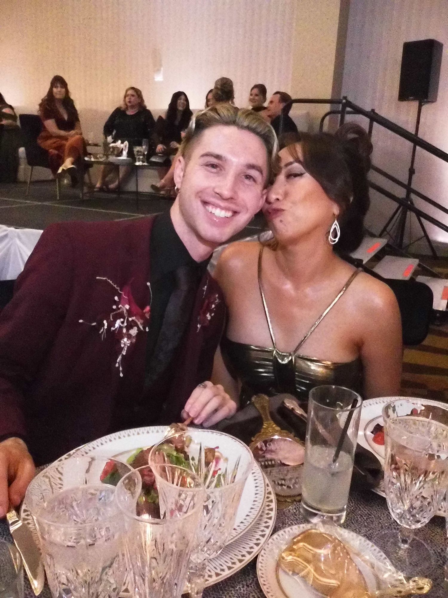Couple posing at formal dinner table, man in burgundy suit, woman in black dress making kissing face.