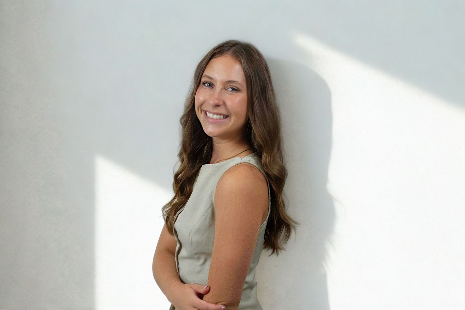 Young woman with long brown hair smiling, wearing a sleeveless gray top, posing against a white wall.