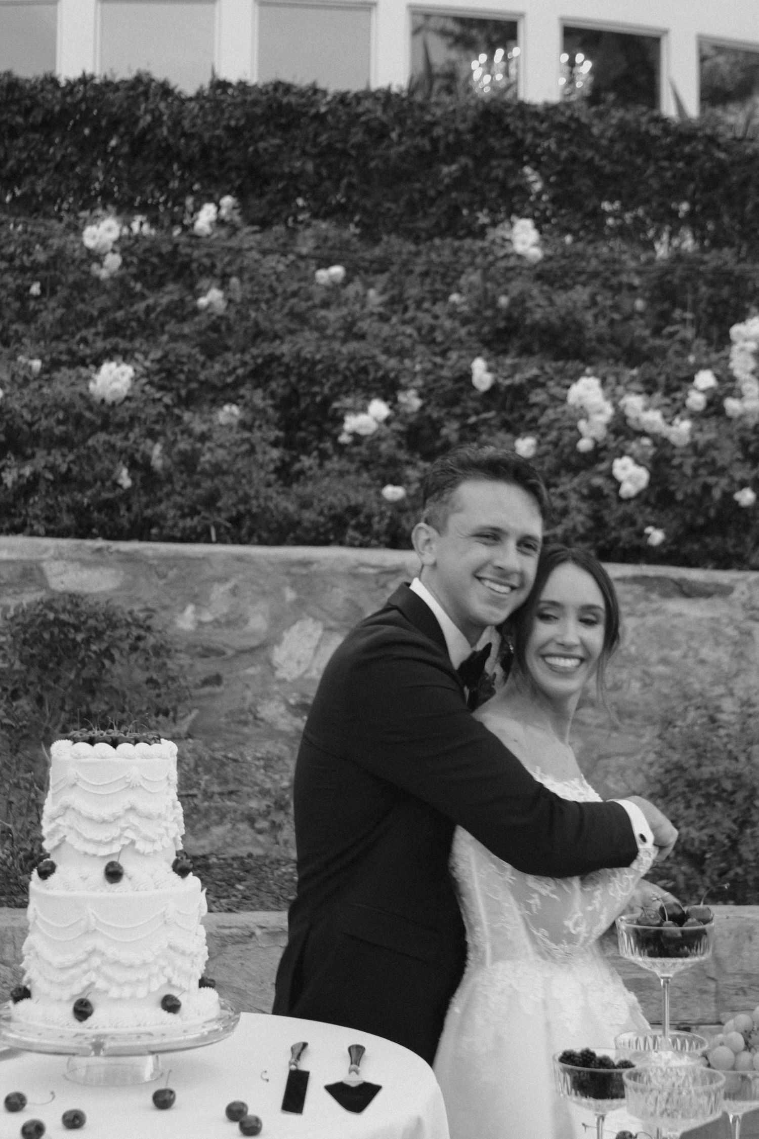 Smiling couple cutting wedding cake outdoors in black and white photo with floral backdrop.