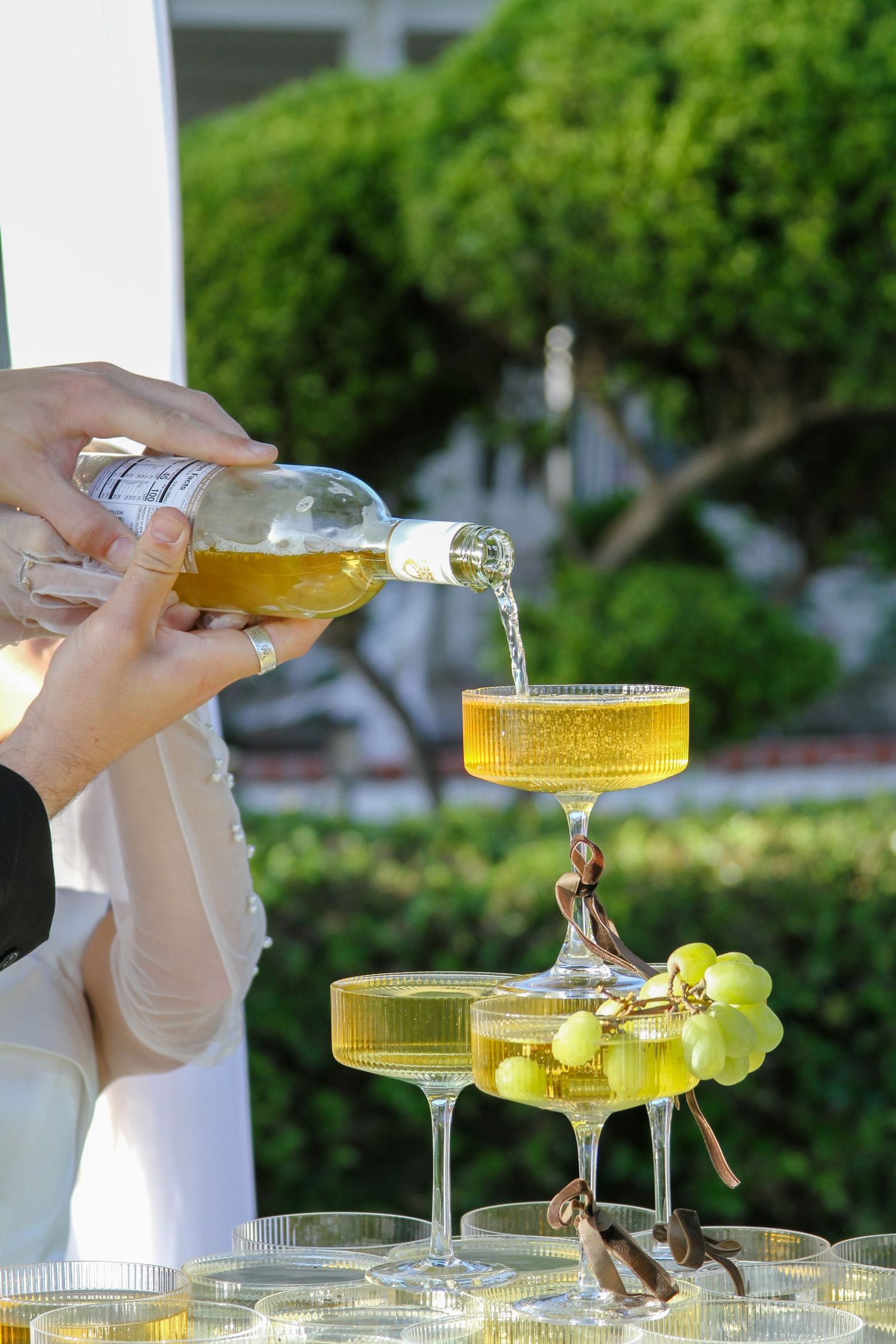 Champagne being poured into a coupe glass tower with green grapes, set outdoors at an elegant event.