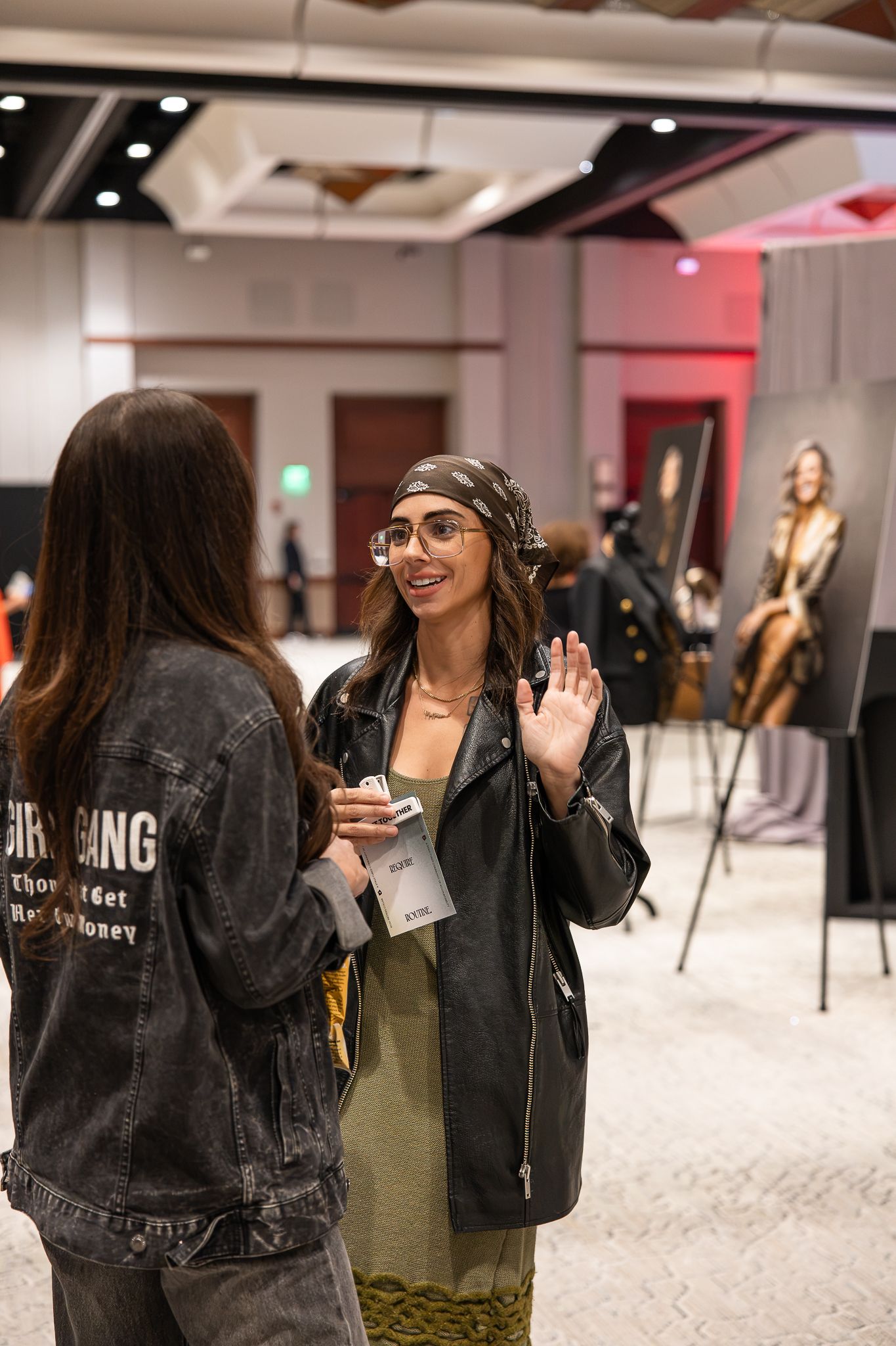 Two women in stylish leather jackets converse at an indoor event, one wearing glasses and a headscarf.