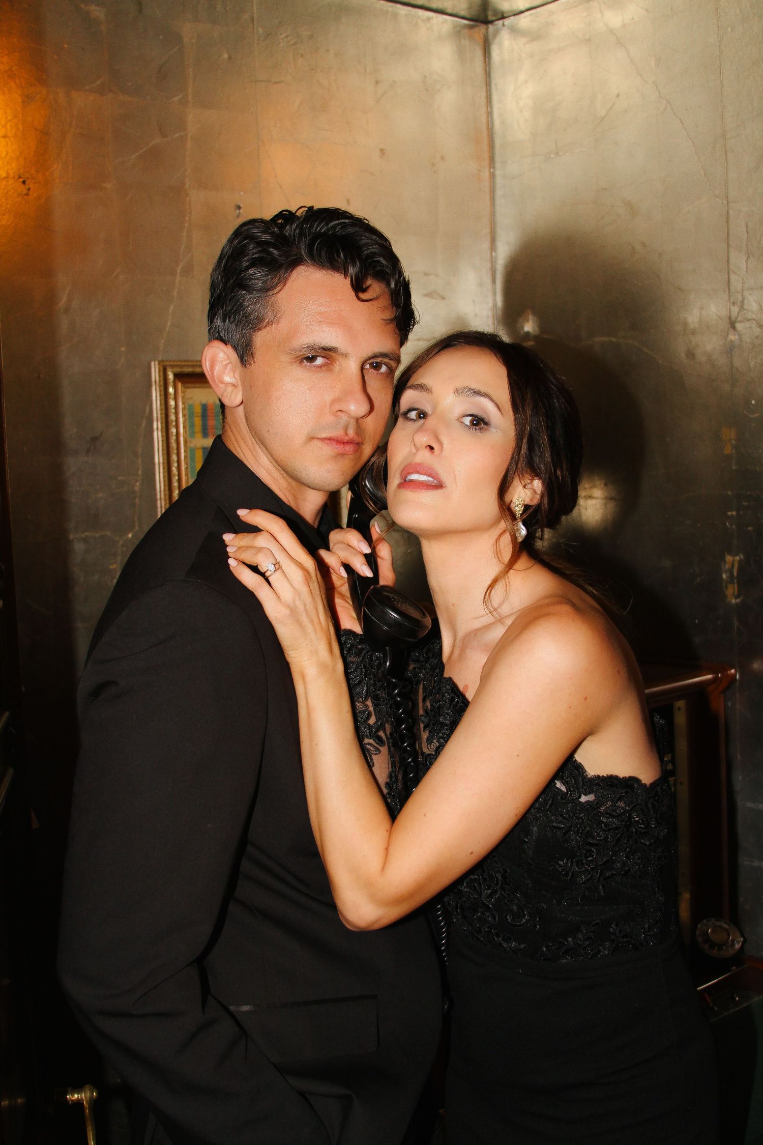 A glamorous couple poses together against an industrial wall in moody lighting.