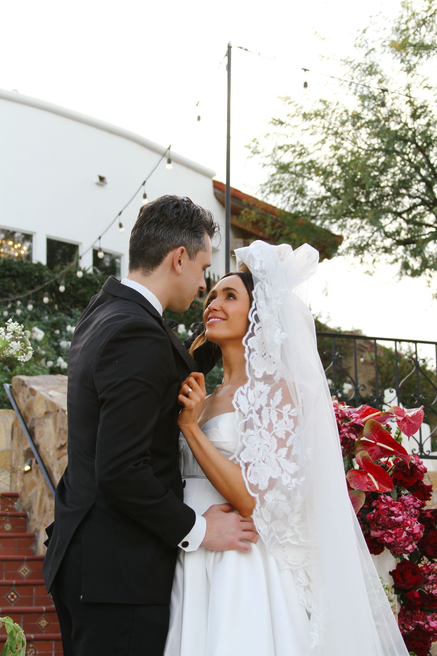 A romantic wedding moment between newlyweds in front of red bougainvillea flowers and stone stairs.