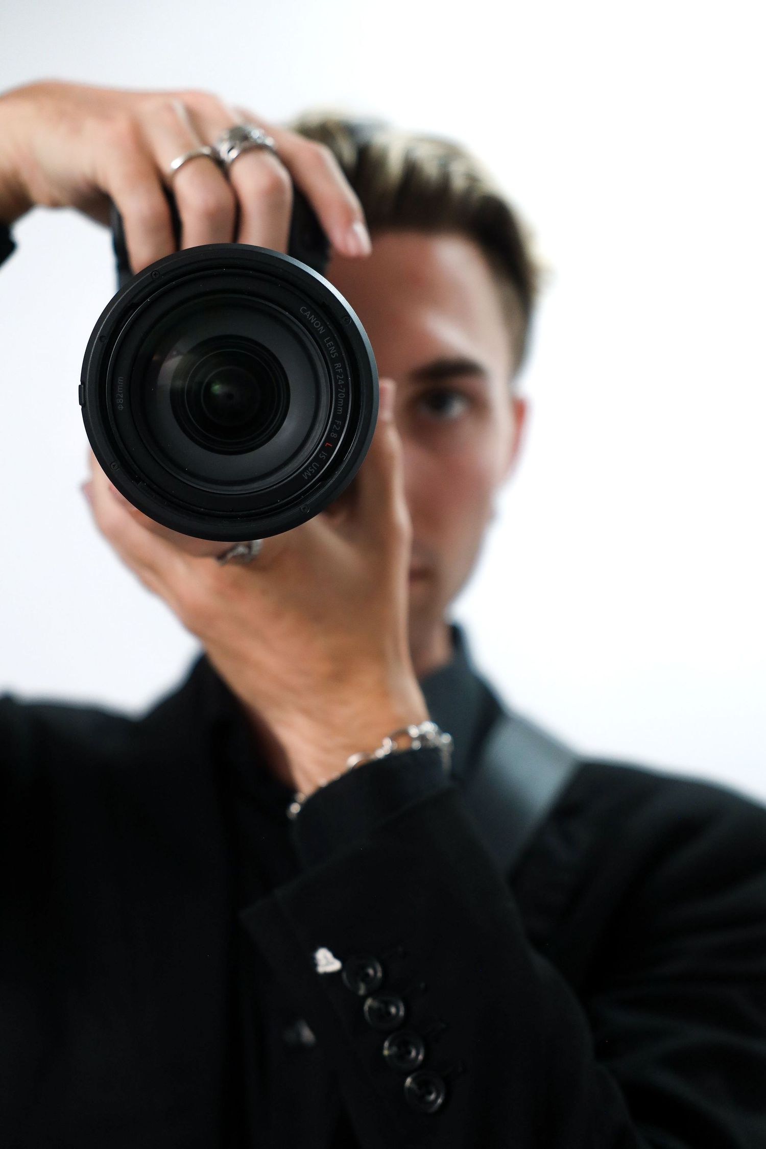 A male photographer in a black suit holds a DSLR camera up to his eye, pointing the lens directly at the viewer.