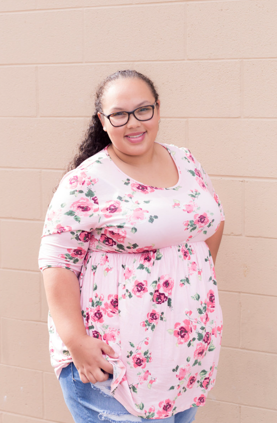 A smiling person wearing glasses and a pink floral blouse with floral print poses against a beige wall.