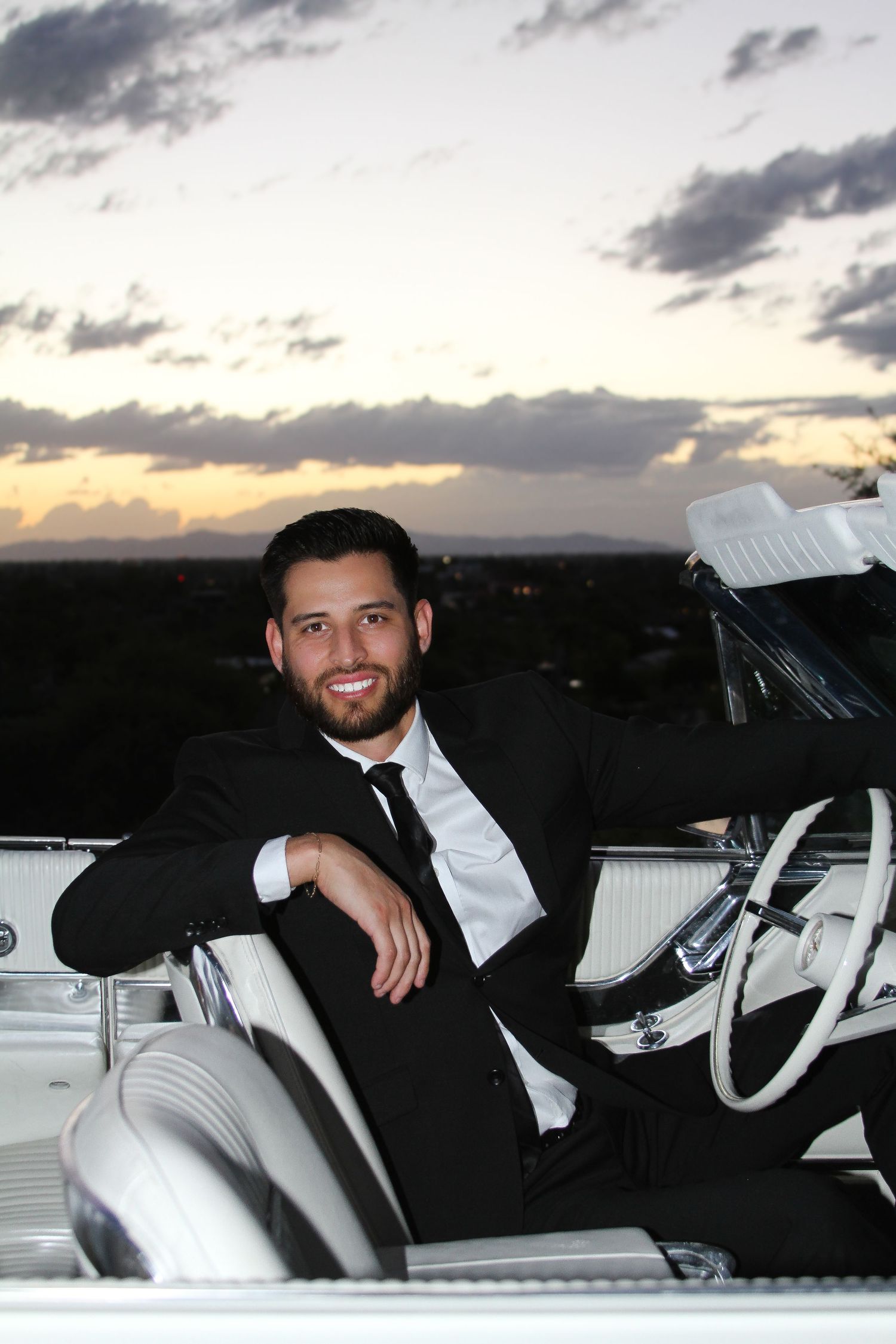 A groom in a black suit sits in the driver's seat of a vintage white car at sunset.